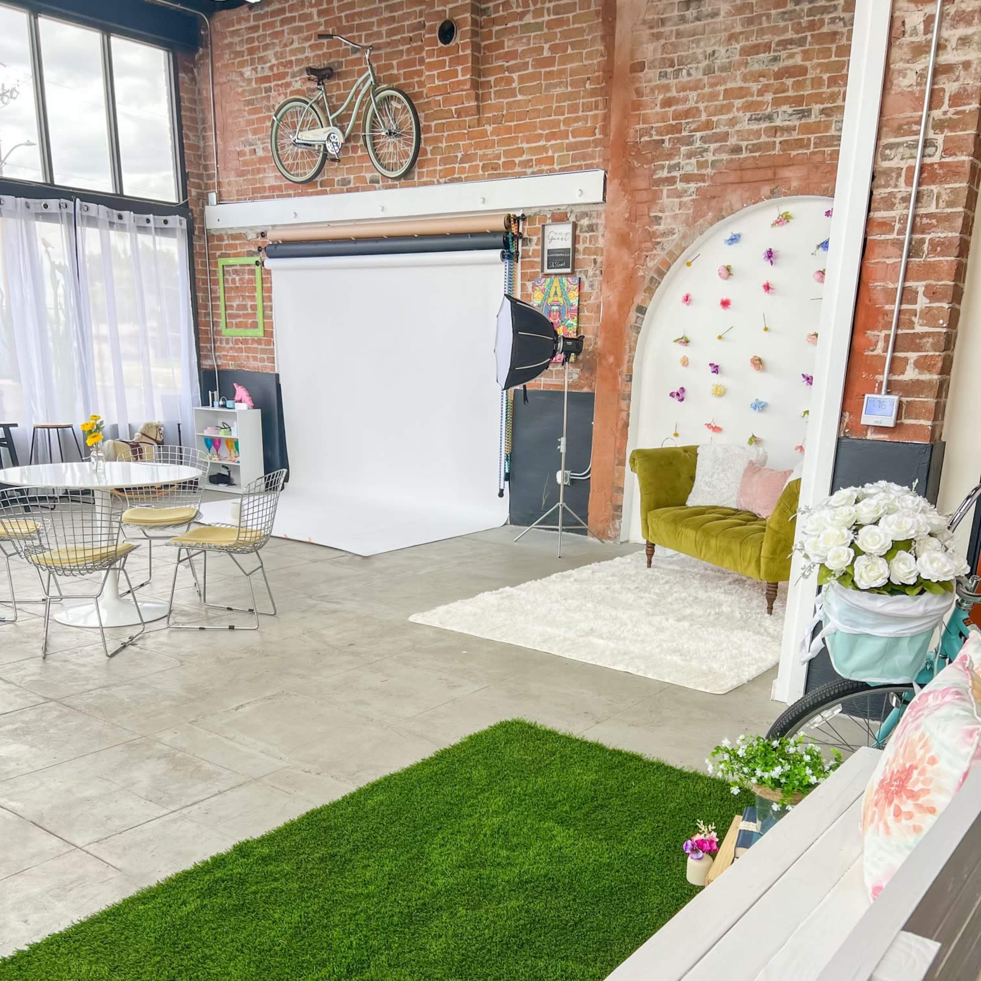 The scene features a photography studio with a white backdrop, a green carpet, seating areas, and decorative elements like flowers and a bicycle on exposed brick walls.