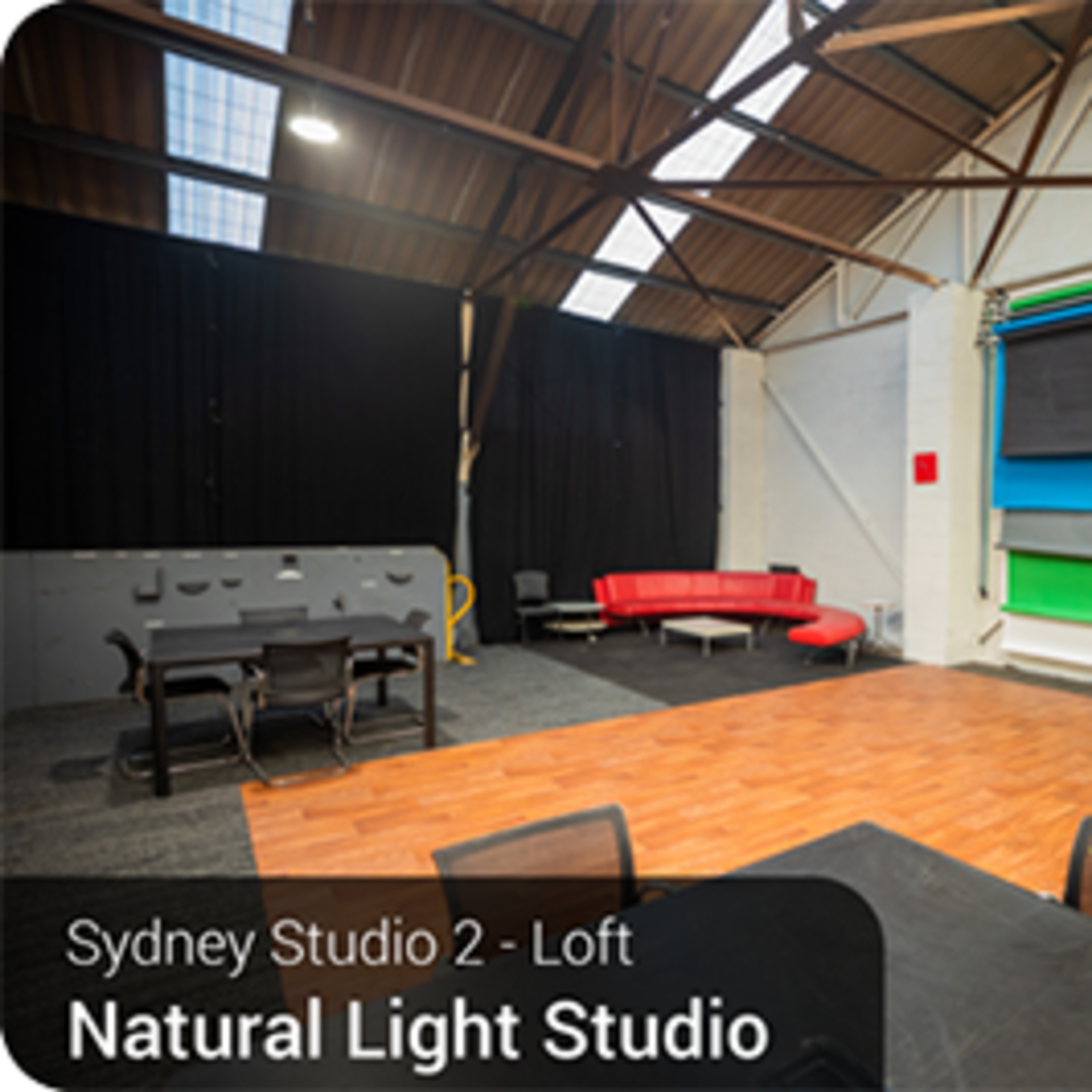 A spacious studio with a wooden floor, featuring a red seating area, several colored backdrops, and a large open ceiling with natural light.