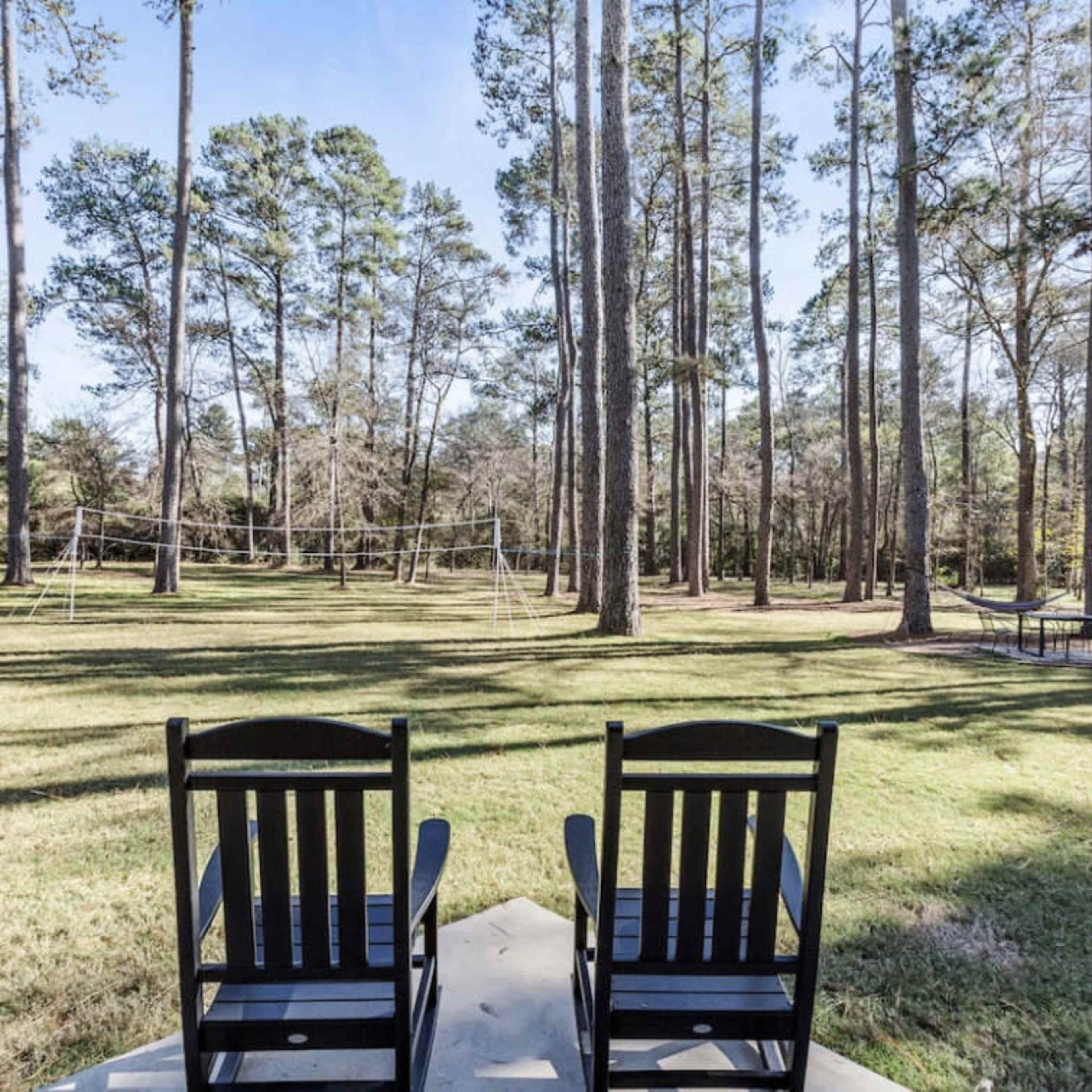 Two black rocking chairs sit on a patio overlooking a grassy area surrounded by tall trees, with a volleyball net in the distance.
