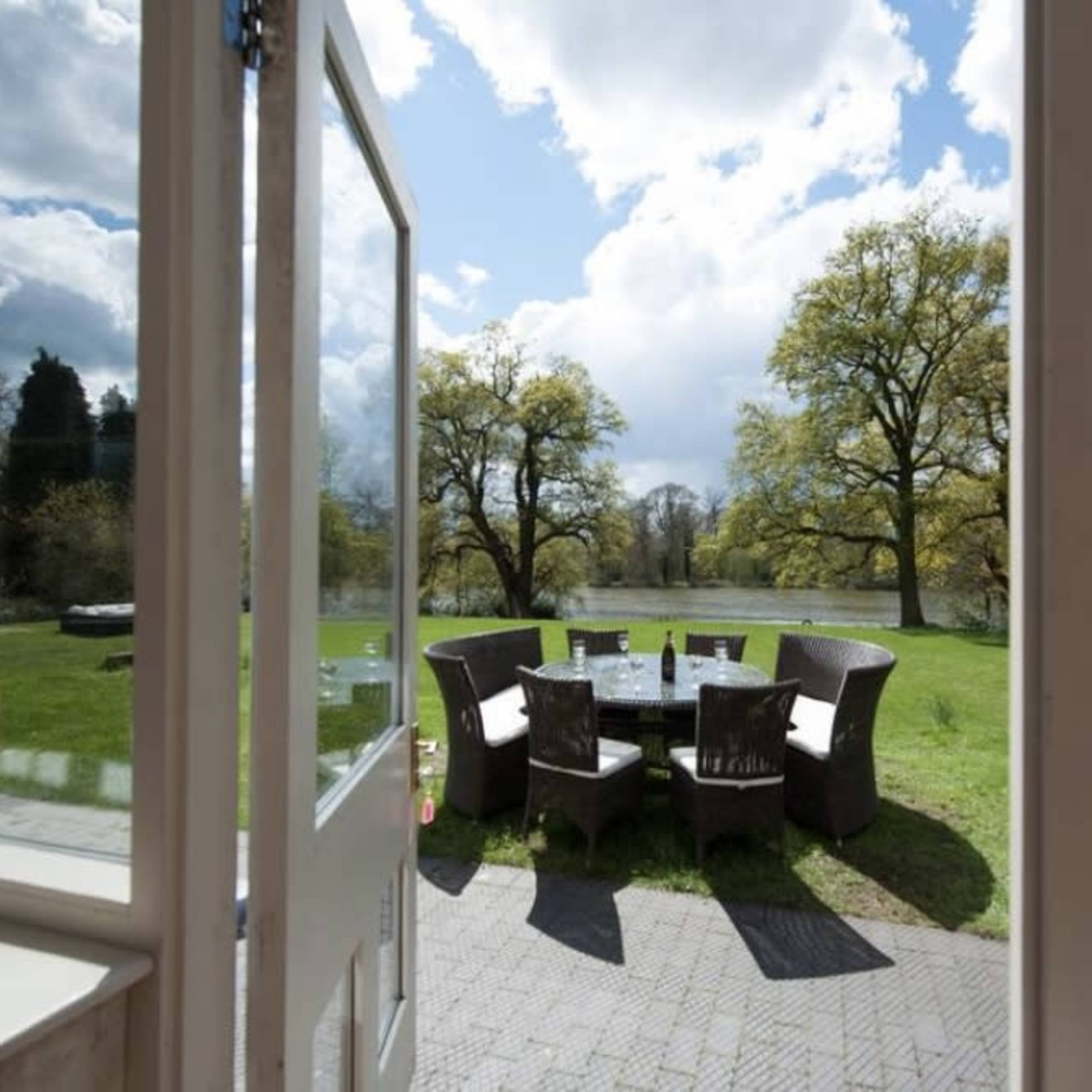 A view from an open door shows a patio with circular seating overlooking a green lawn and trees under a cloudy sky.