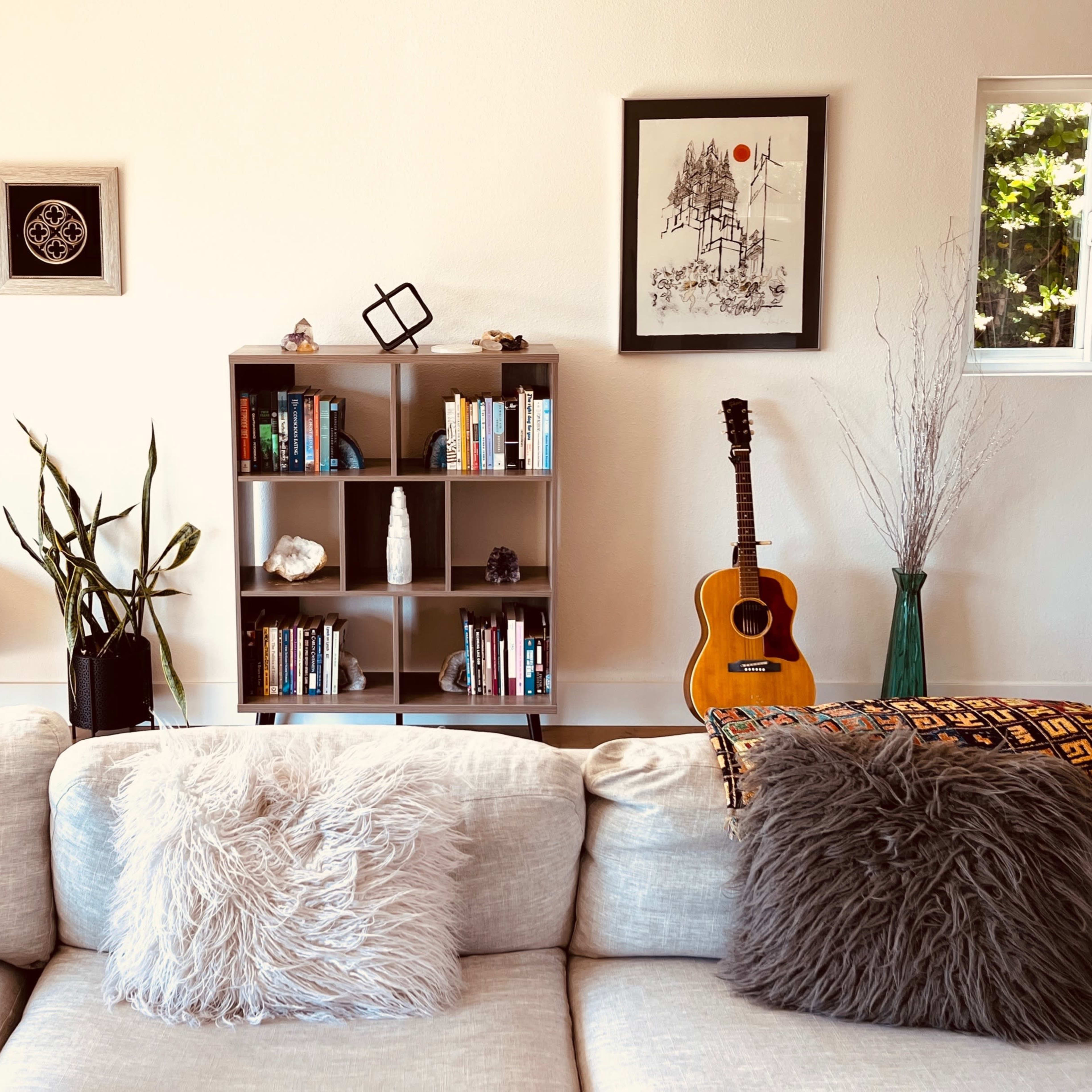 A cozy living room with a beige couch adorned with two fluffy pillows, a bookshelf filled with books, and various decorative items, including a guitar and a nearby potted plant.