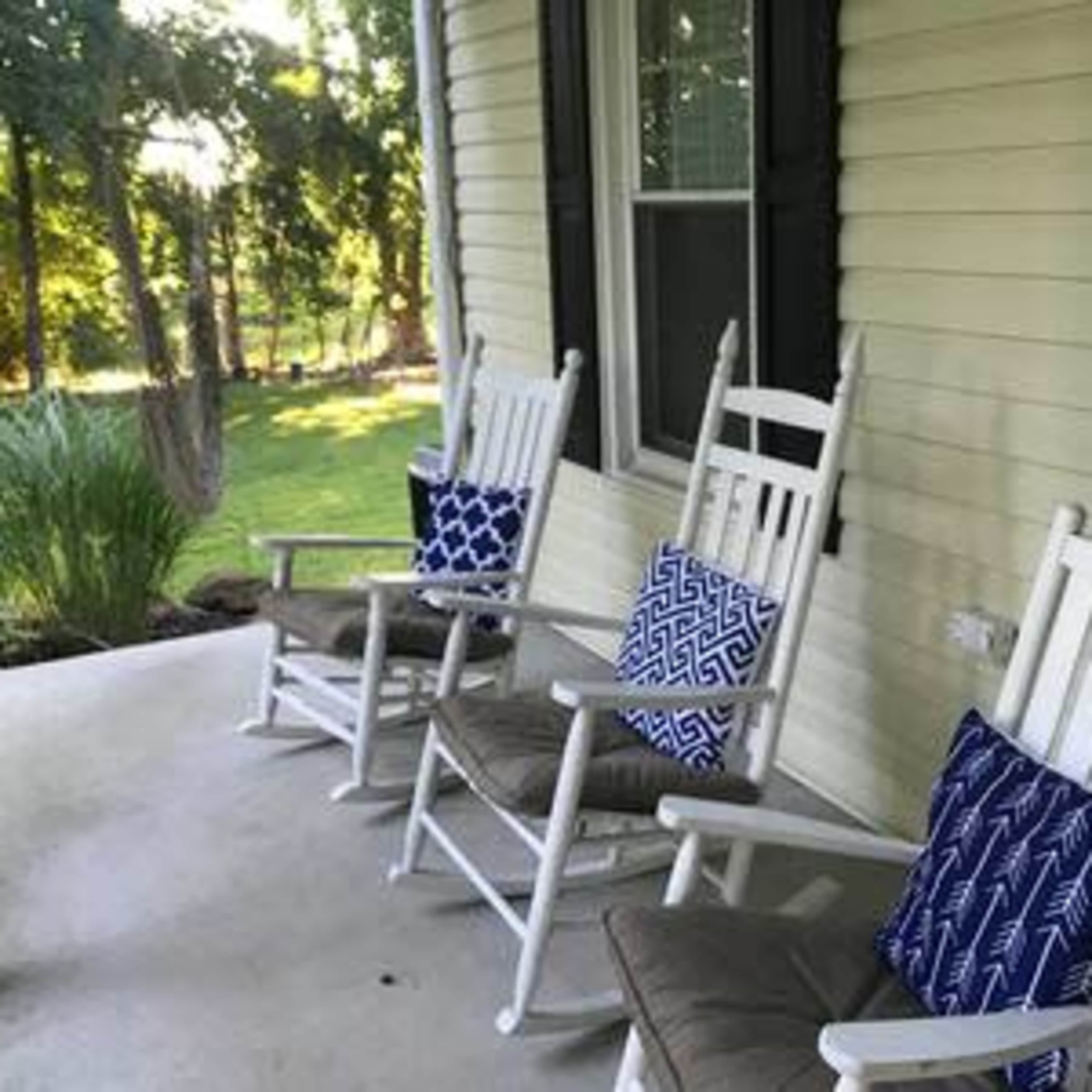 A porch with three white rocking chairs, each adorned with blue patterned cushions, overlooking a green yard.