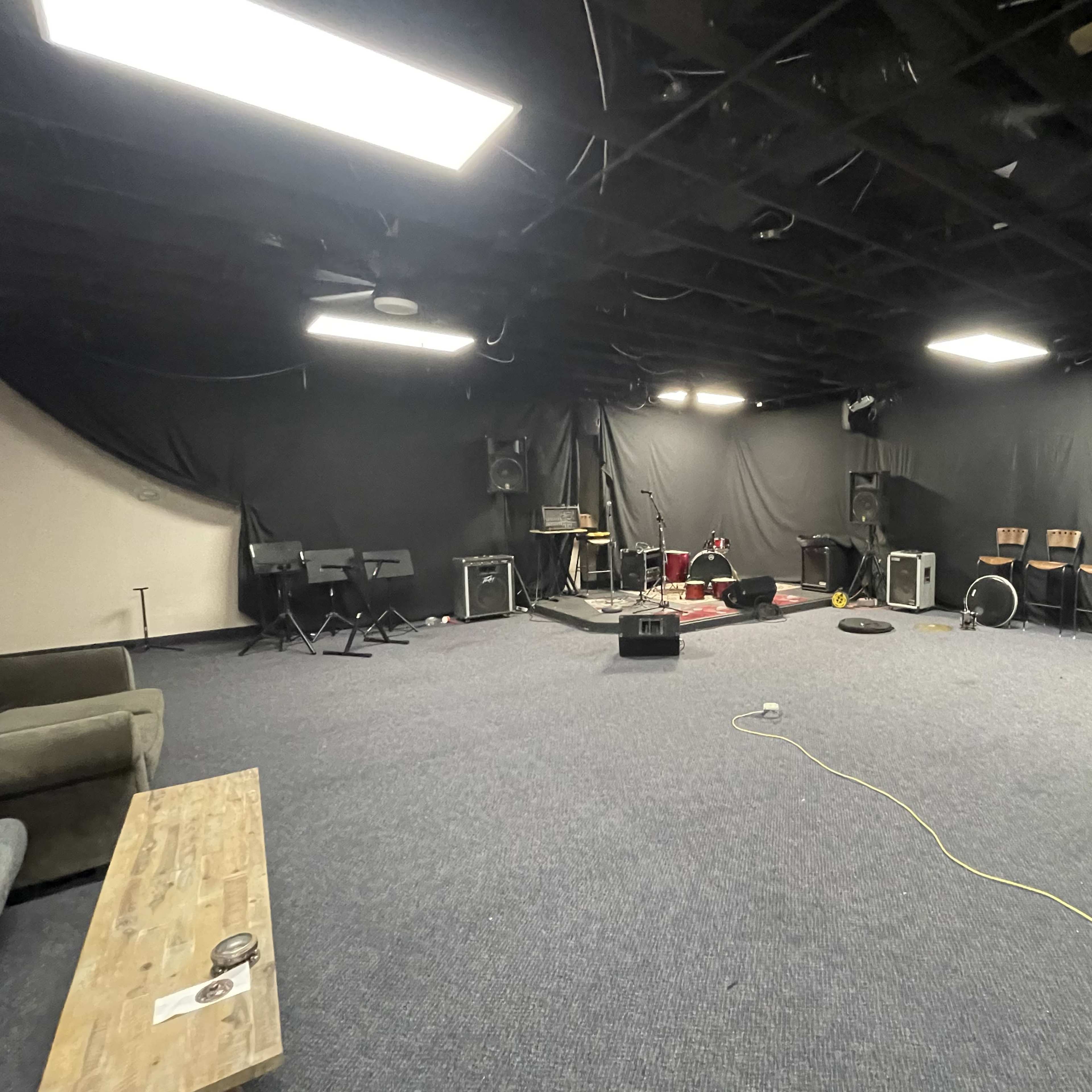 A large, empty rehearsal space with black walls, several musical instruments set up in the center, and seating along the perimeter.