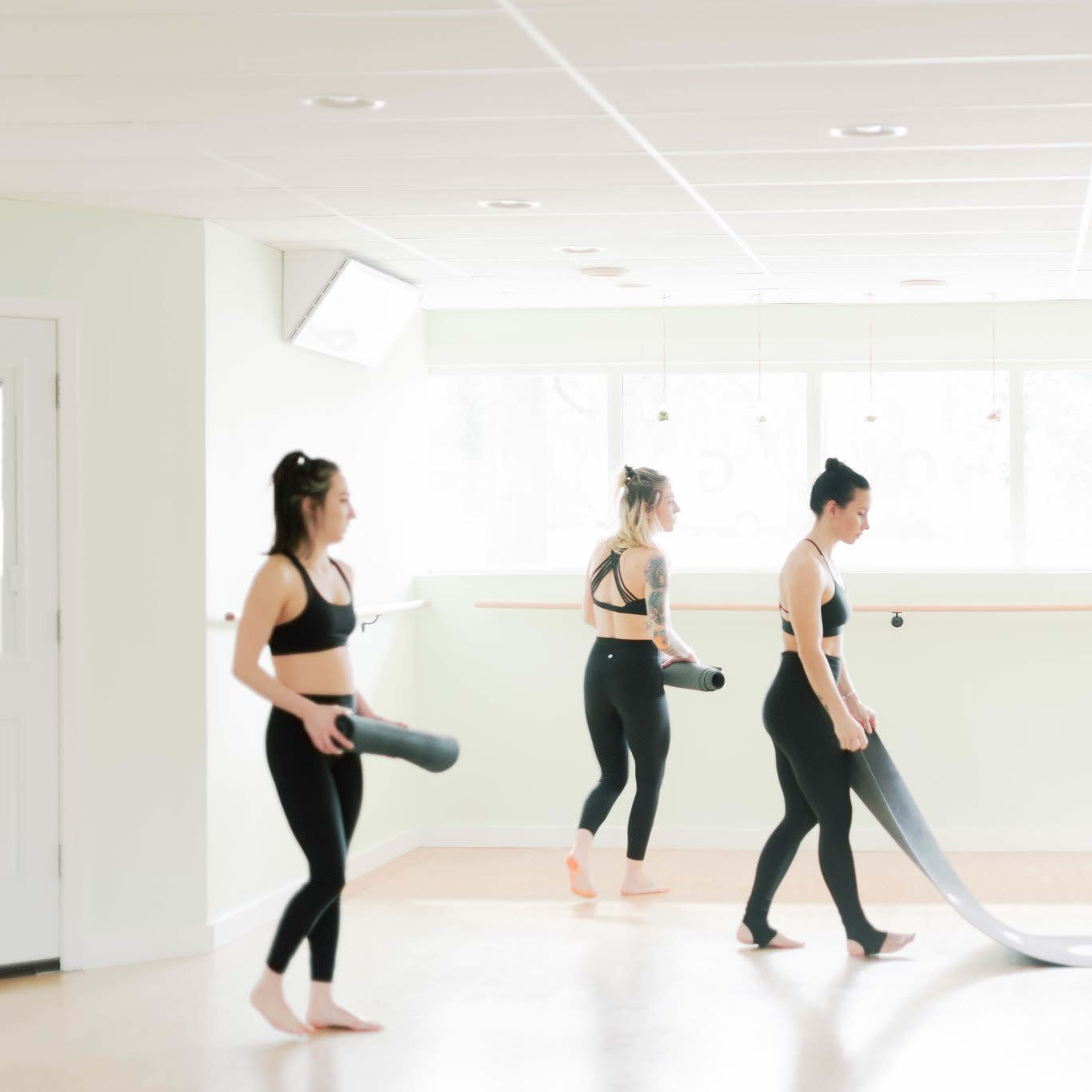 Three women in athletic clothing are rolling out yoga mats in a bright, minimalistic studio with large windows.