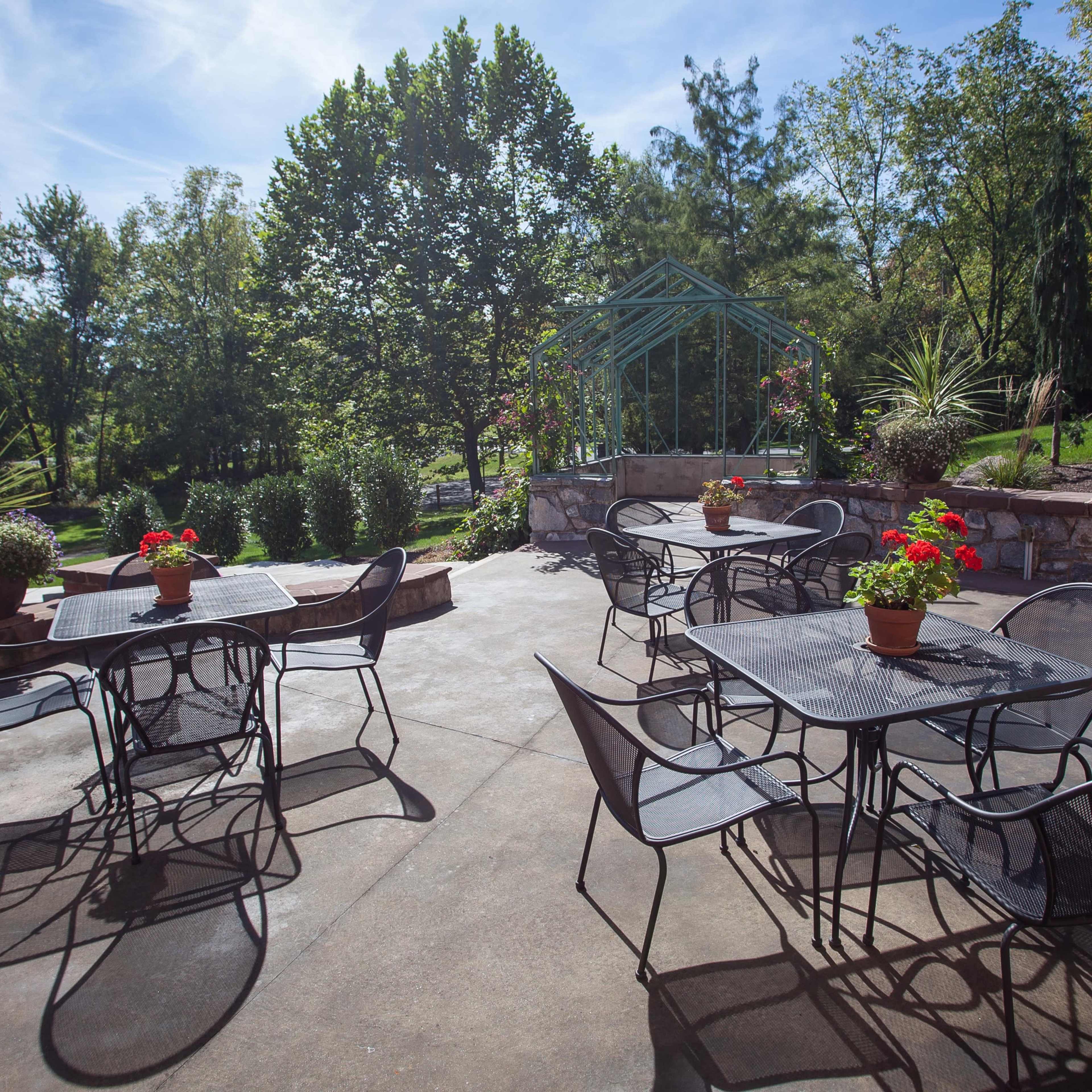 A patio area features several metal tables and chairs arranged with potted plants, surrounded by greenery and trees.