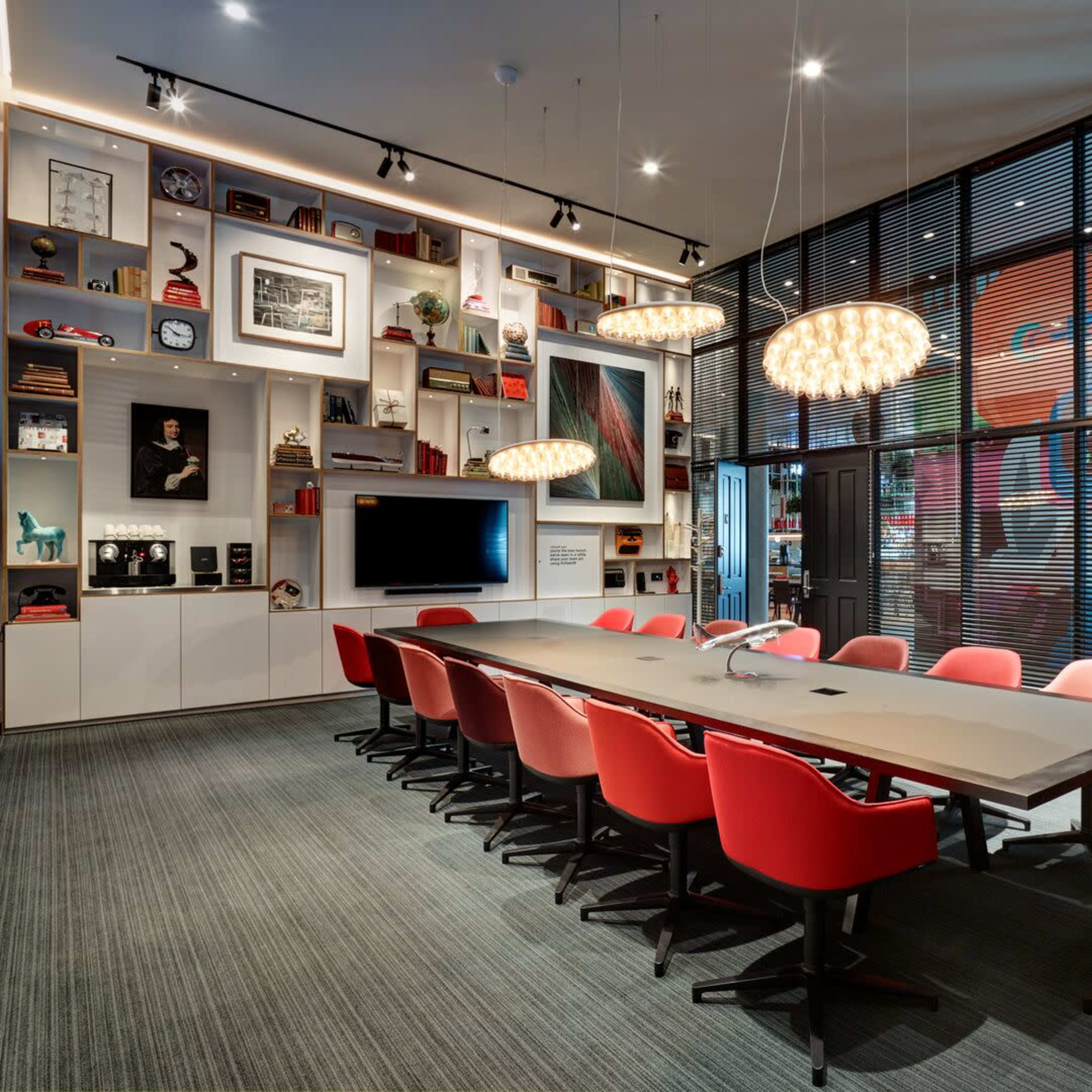 A modern conference room features a long table surrounded by red chairs, with a wall of shelves displaying art and decor, and large windows overlooking a colorful mural.