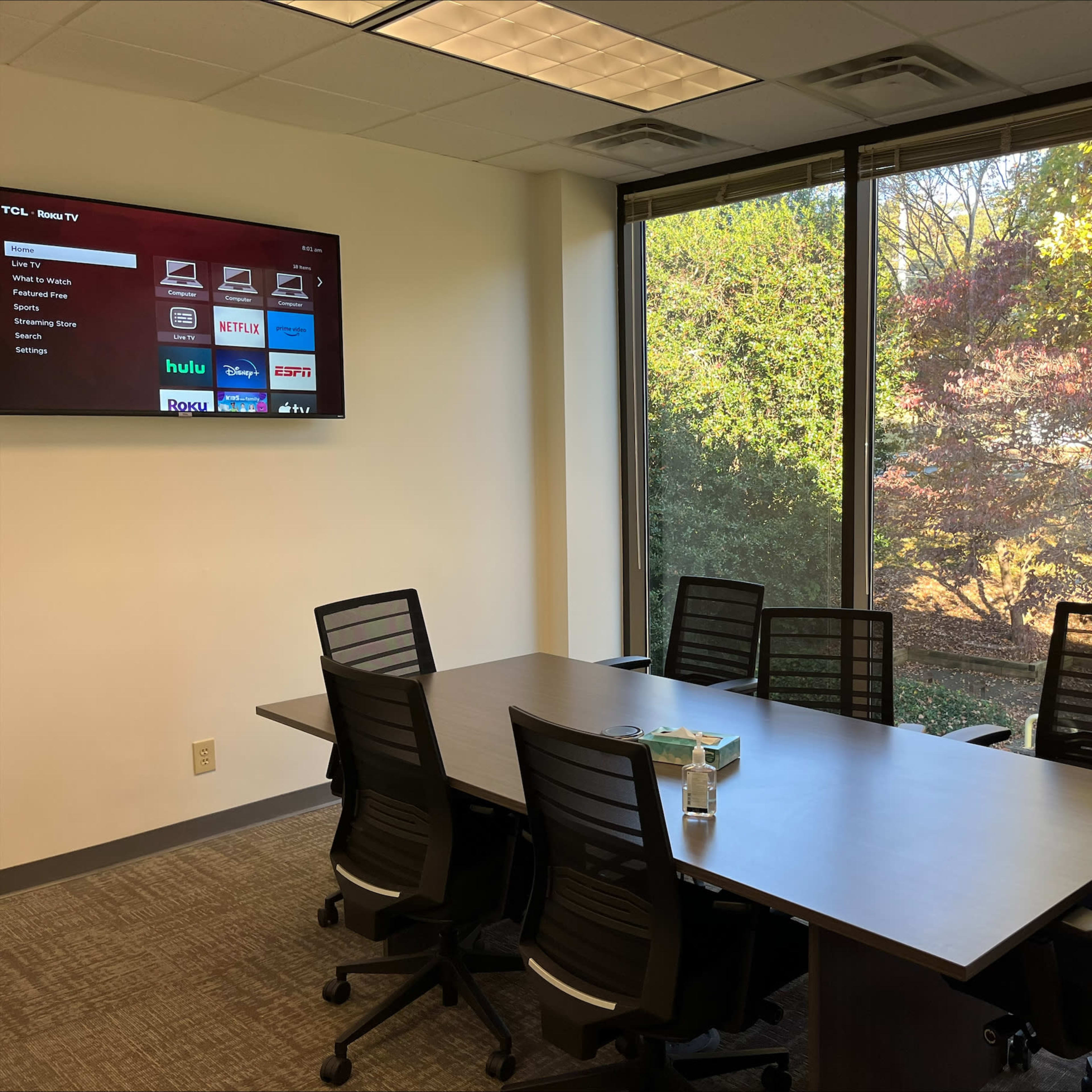 A conference room features a long table surrounded by ergonomic chairs and a wall-mounted screen displaying various streaming service logos.
