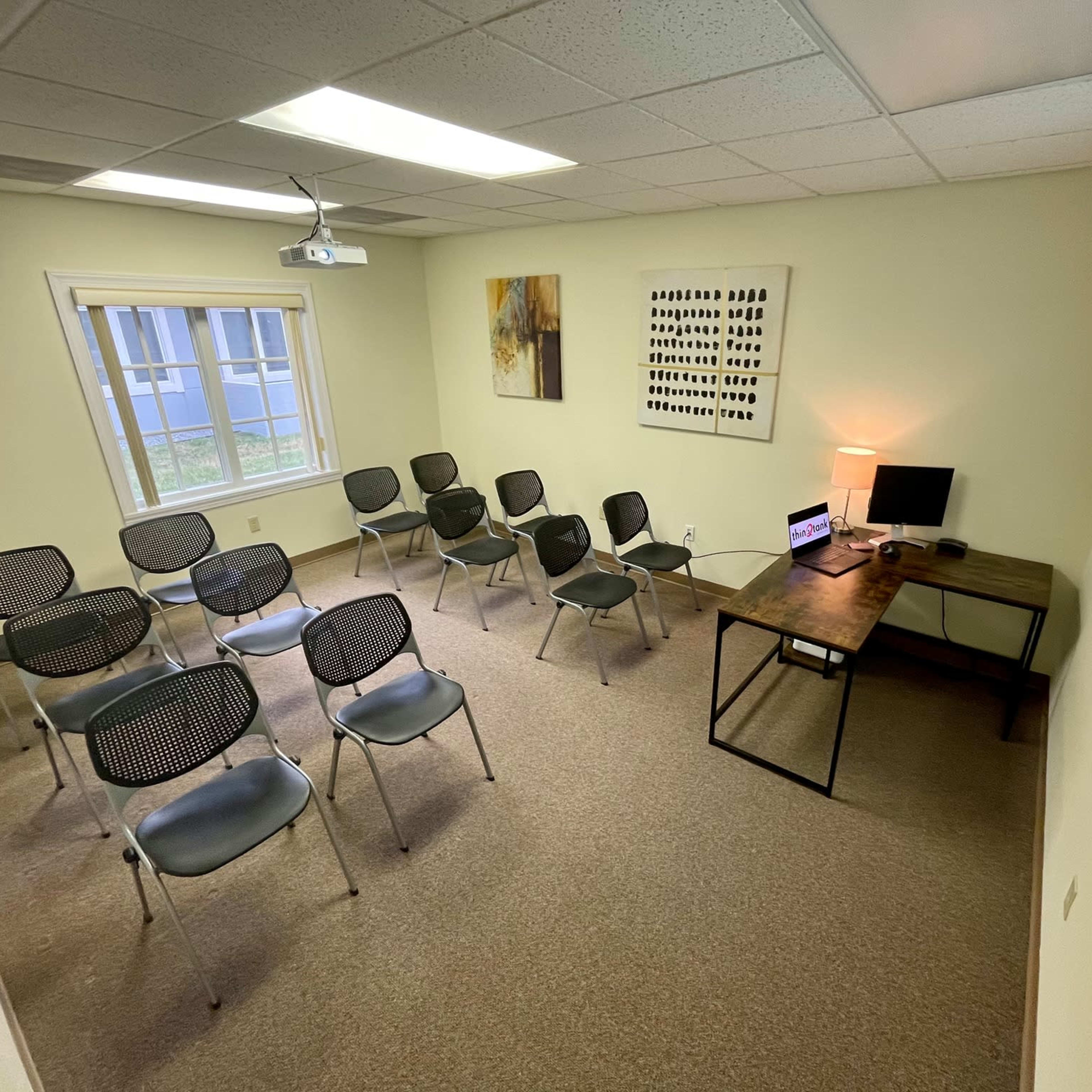A small meeting room with seated chairs arranged in rows, a desk with a laptop, and a wall-mounted projector displaying a logo.