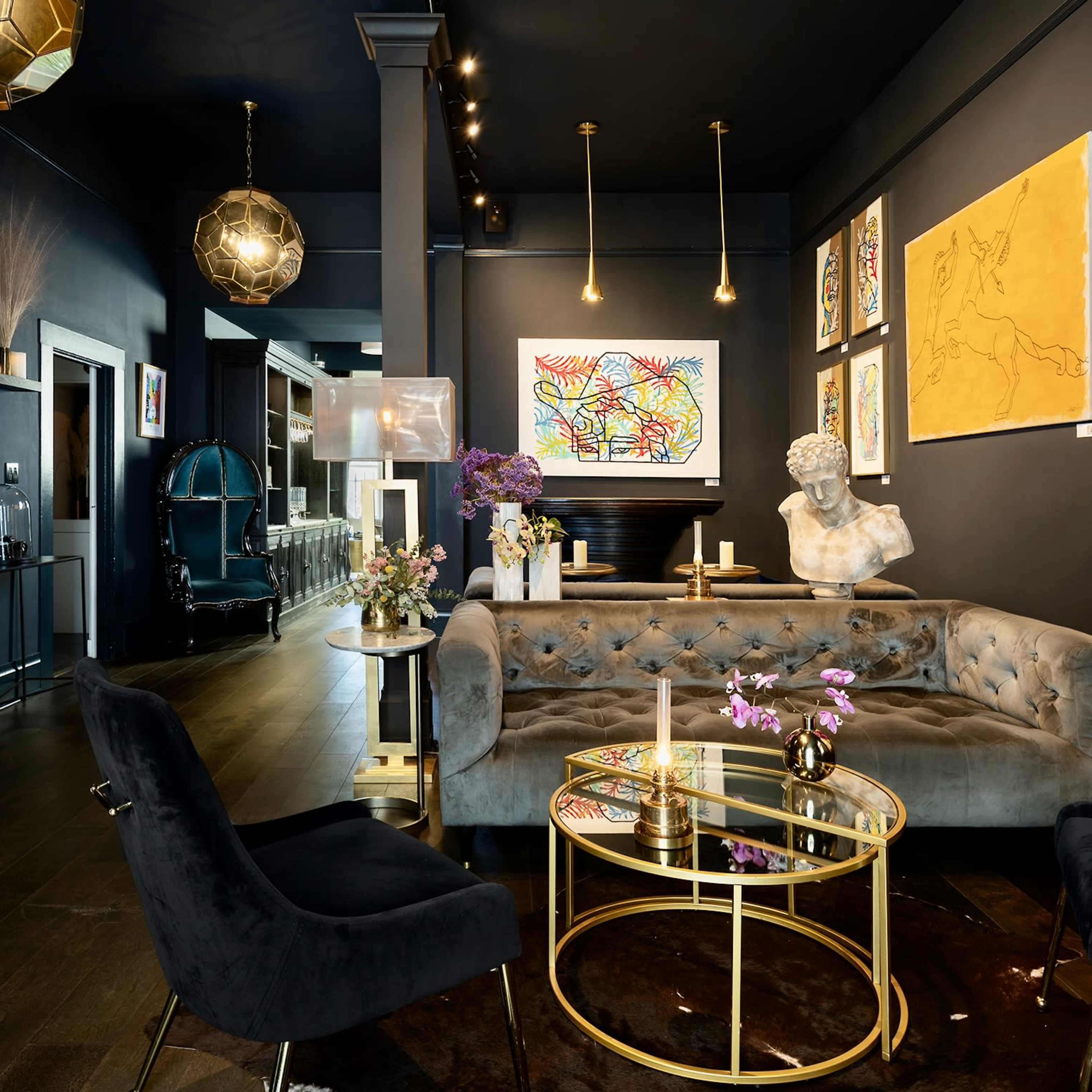 A modern lounge features a gray velvet sofa, a glass coffee table, and colorful artworks on dark walls, with stylish lighting fixtures above.