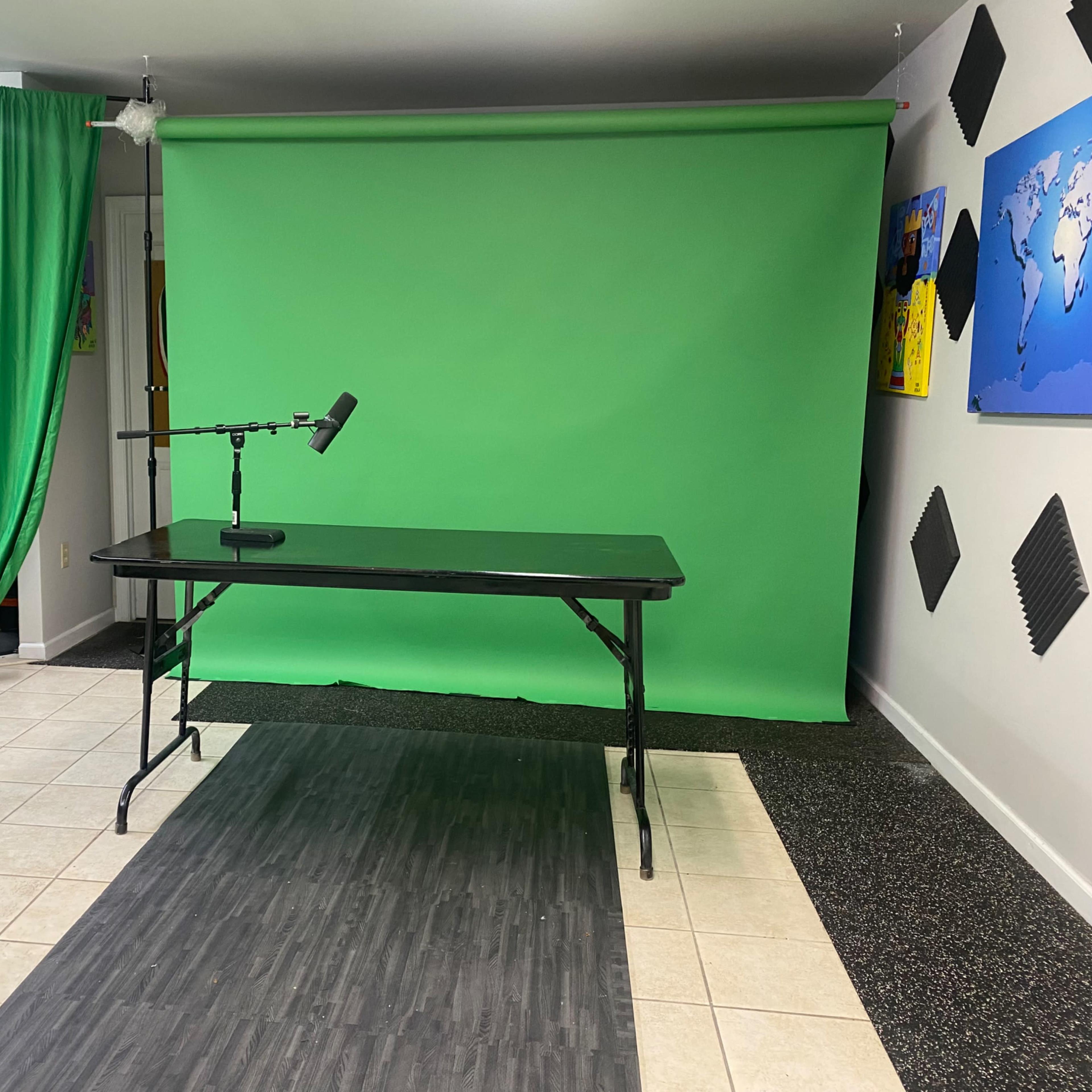 A room set up for video production, featuring a green screen, a black tabletop, a microphone on a stand, and green curtains.