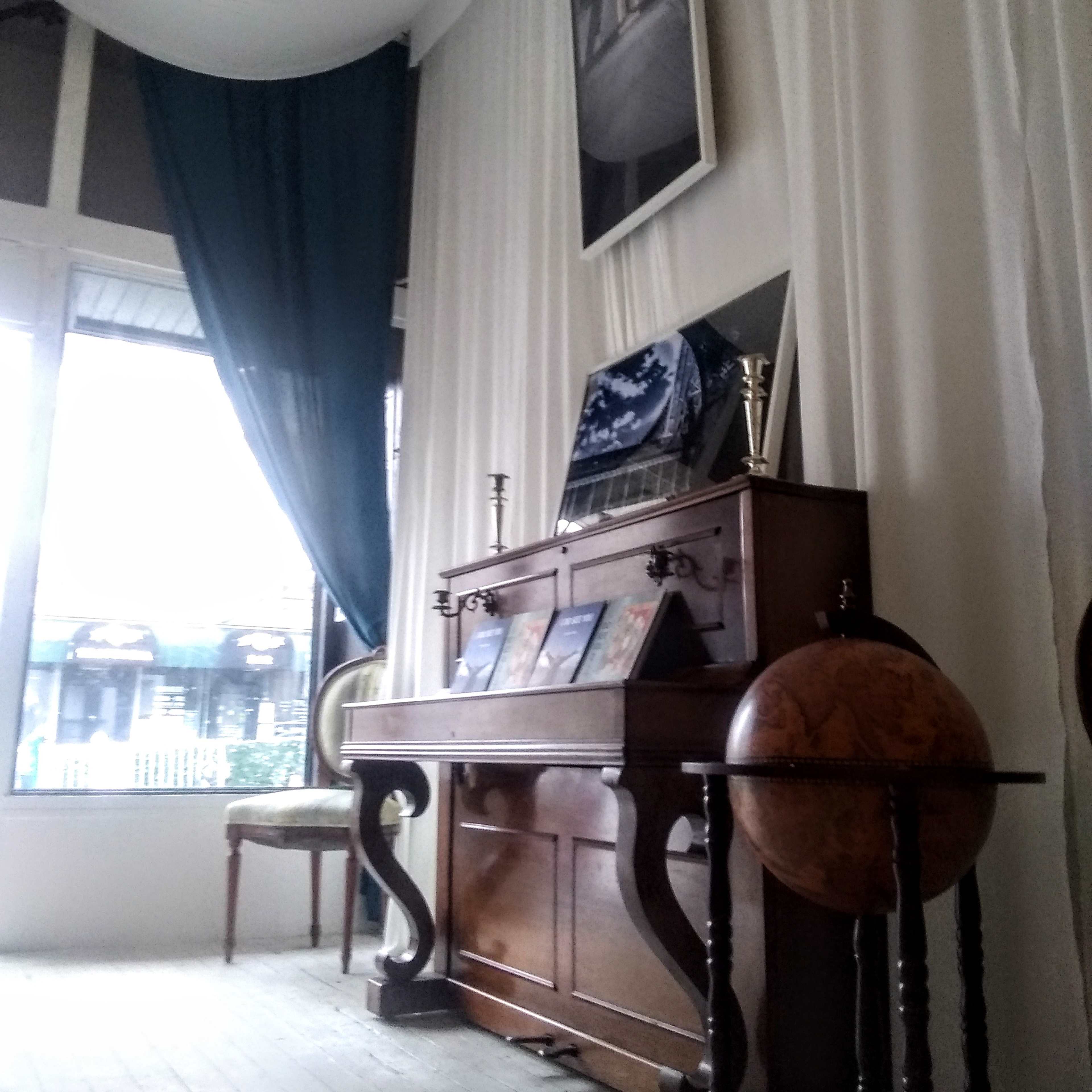 A room with white draped curtains, a wooden piano displaying framed artwork, and a globe on a stand beside a window.