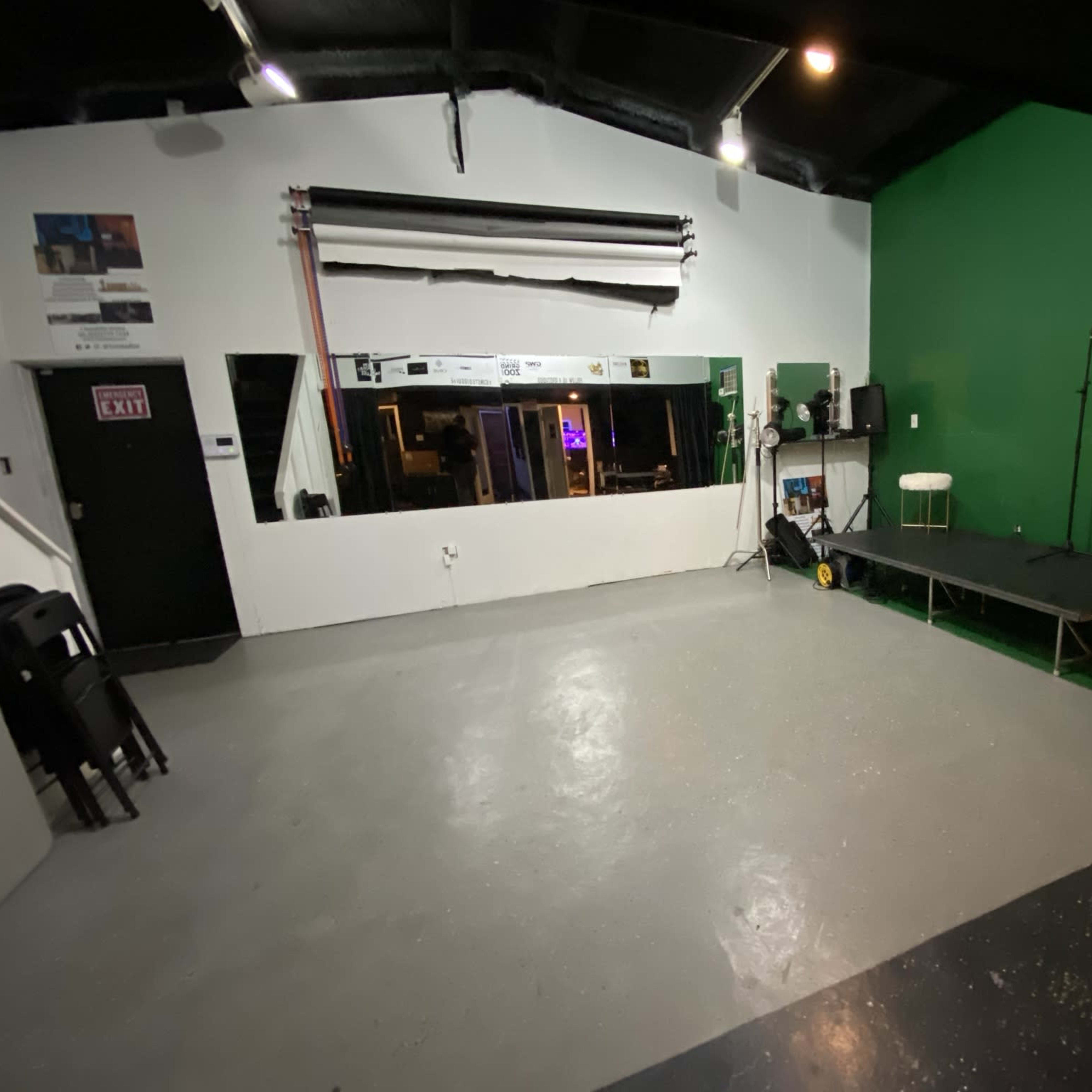 A performance space featuring a green wall, a small stage, audio equipment, and a large mirror opposite the stage.
