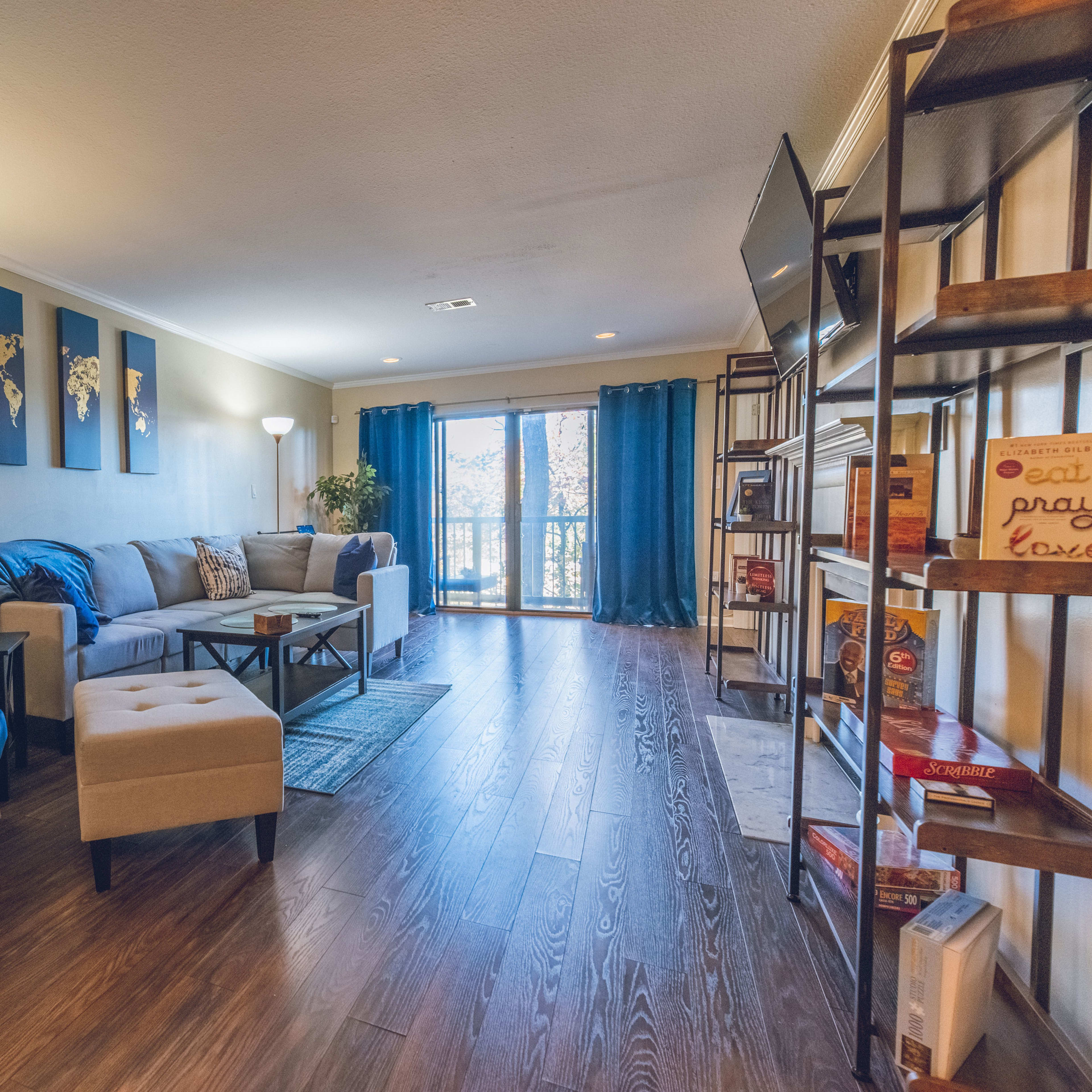 The living room features a couch, an armchair, a coffee table, and a large window with blue curtains, surrounded by bookshelves and decorative items.