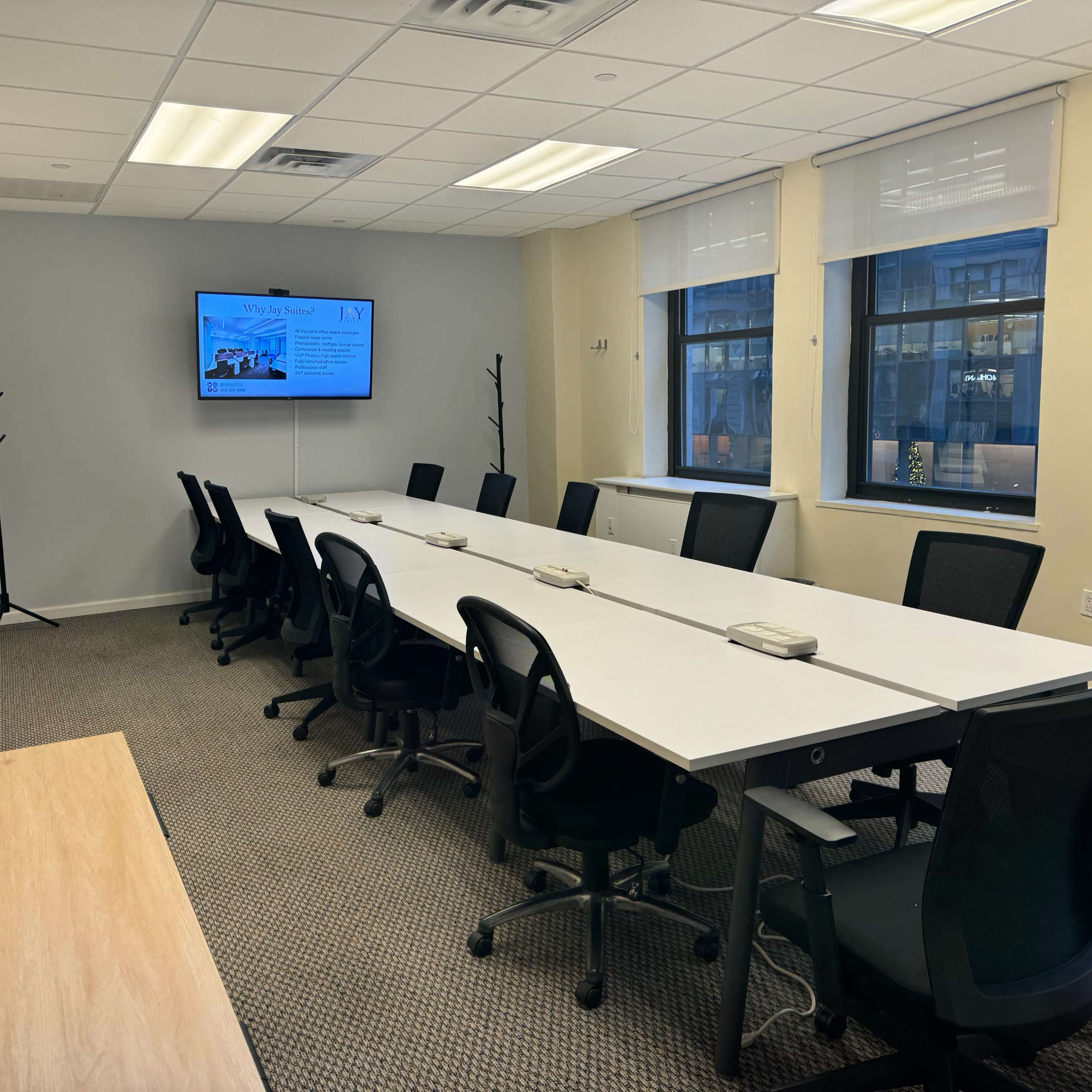 A conference room features a long table surrounded by black chairs, with a television screen displaying information on the wall.