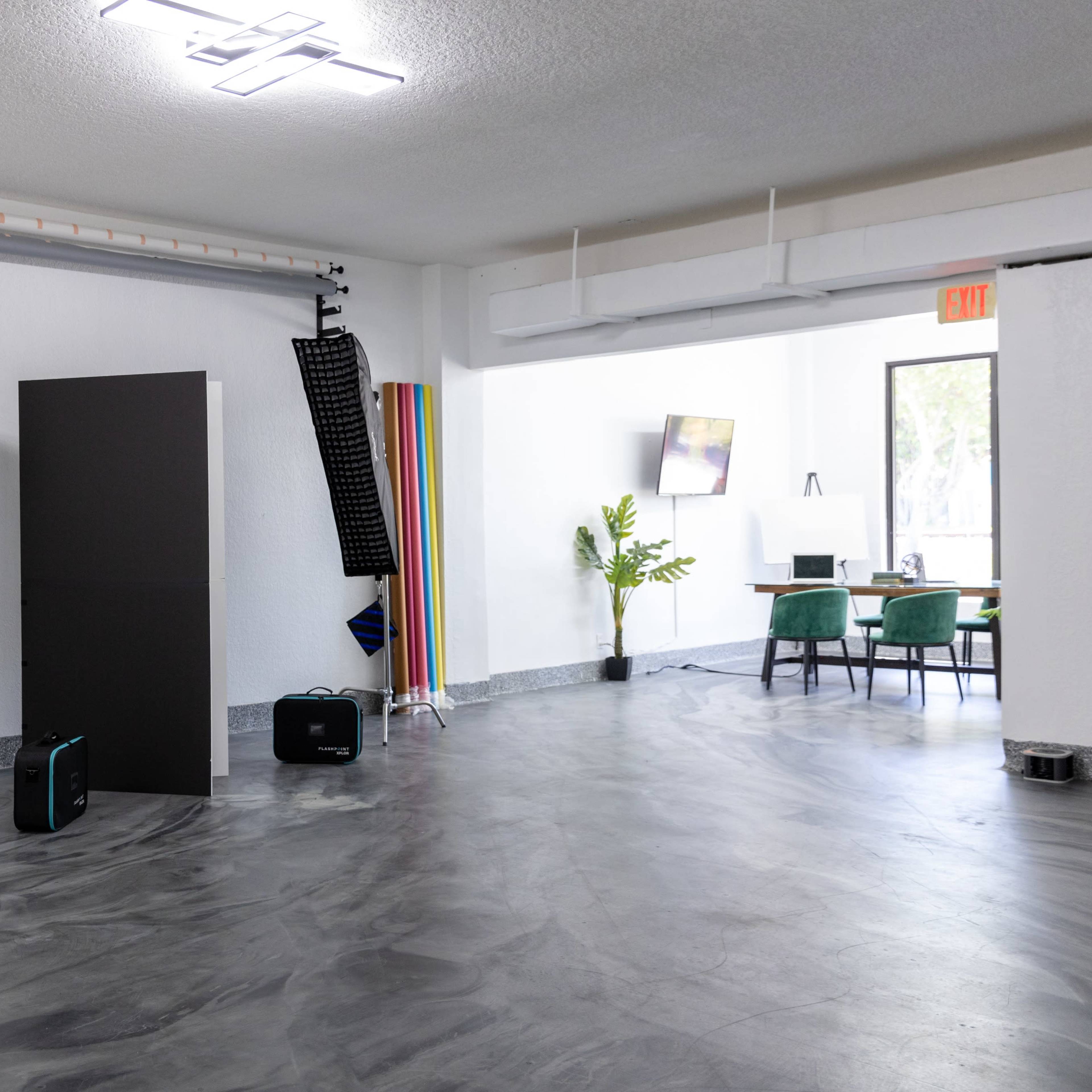 A well-lit studio space with photography equipment, including an umbrella softbox, a black backdrop, and a variety of colored boards, alongside a workspace with a table and chairs.