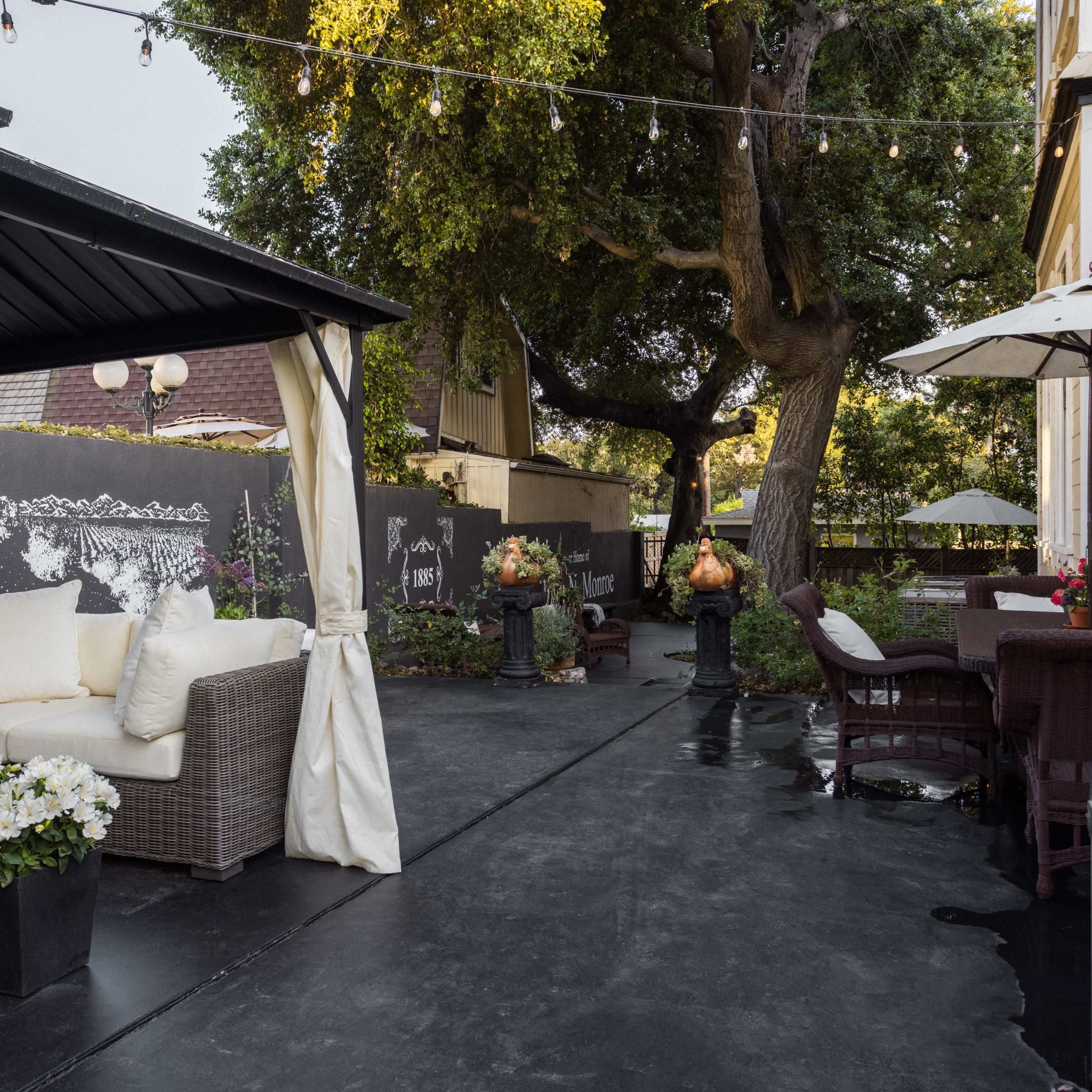 An outdoor patio area with a cozy seating arrangement, decorative plants, and string lights overhead, adjacent to a house and a large tree.