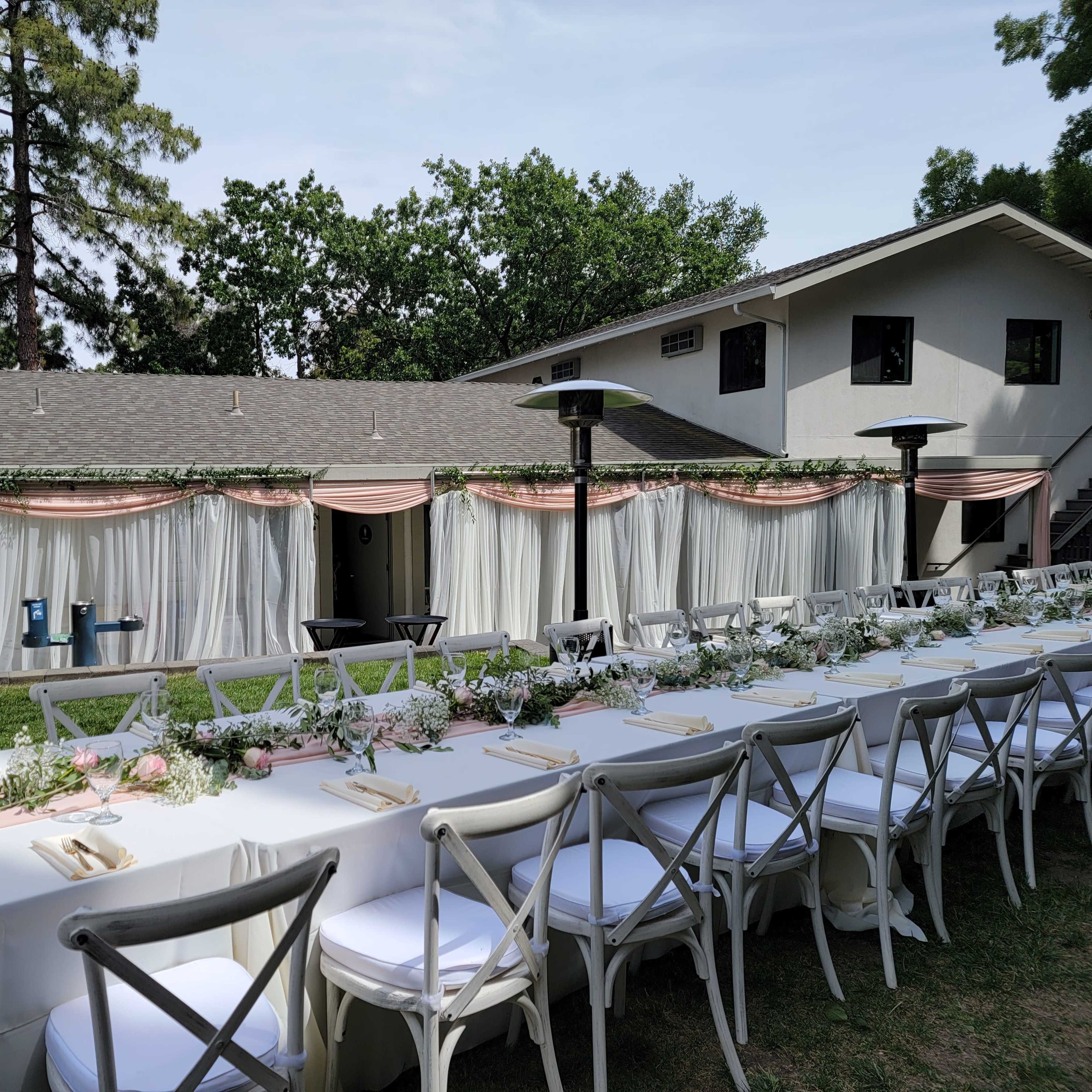 A long, elegantly set dining table with white tablecloths is arranged outdoors, adjacent to a building with draped white curtains and surrounded by greenery.