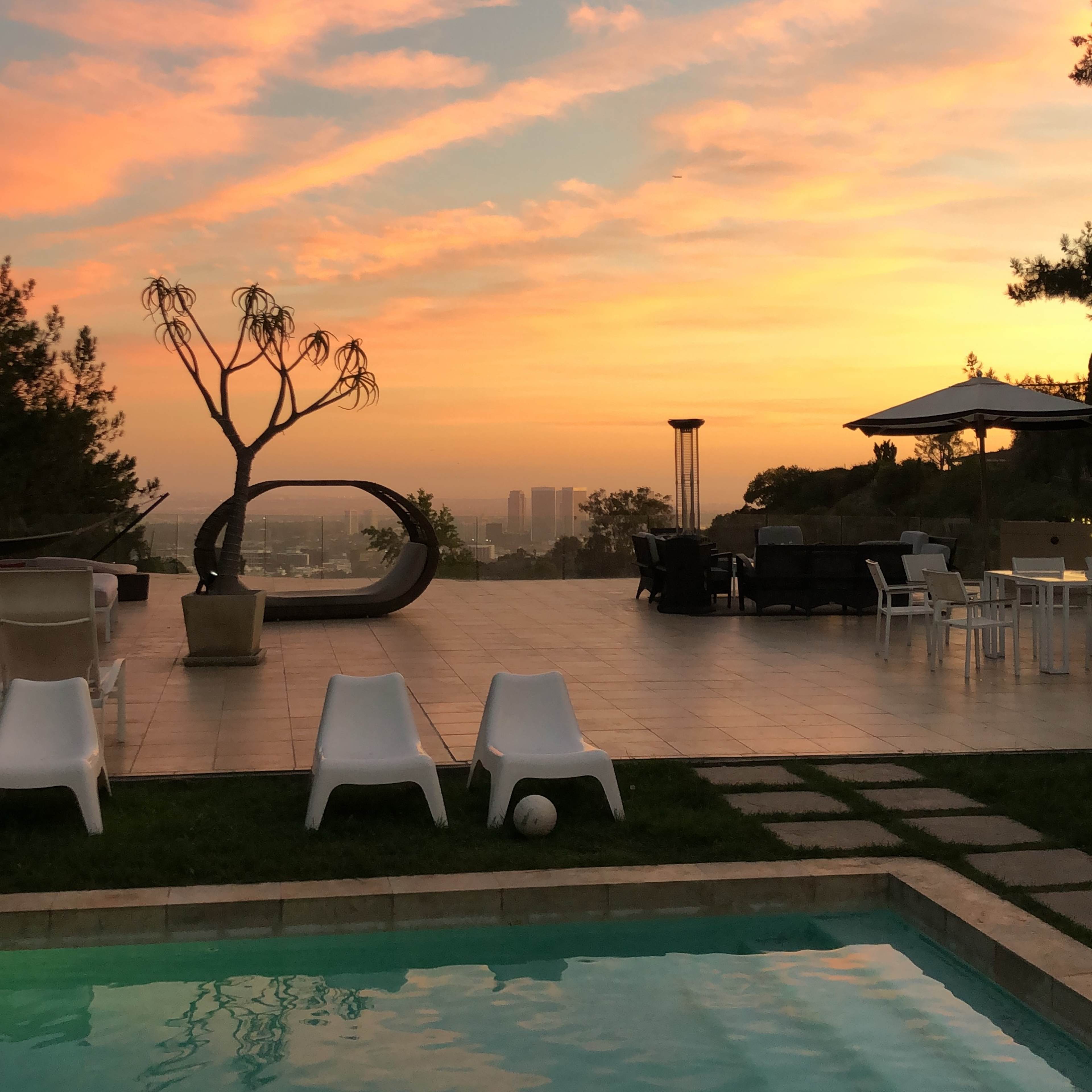 The scene shows a swimming pool surrounded by white chairs and a sunset sky with colorful clouds over a cityscape.