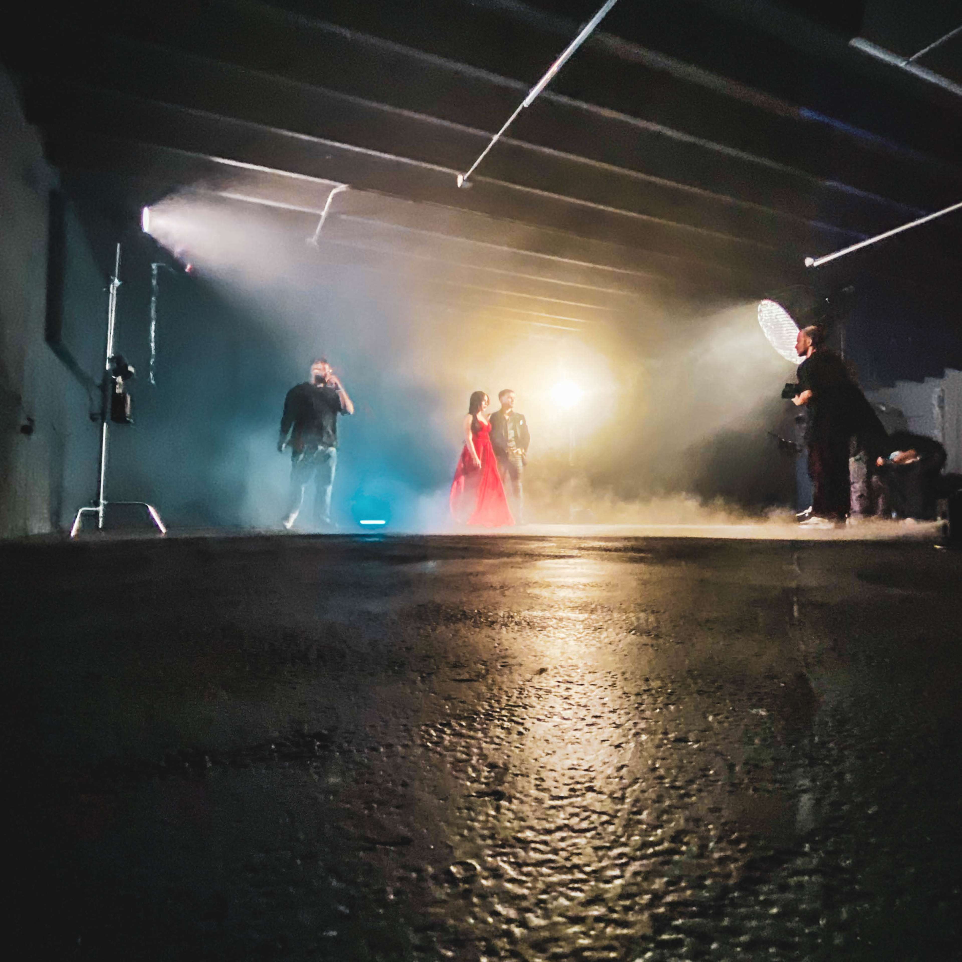 A woman in a red dress stands in a dimly lit warehouse, surrounded by several people and soft lighting effects.