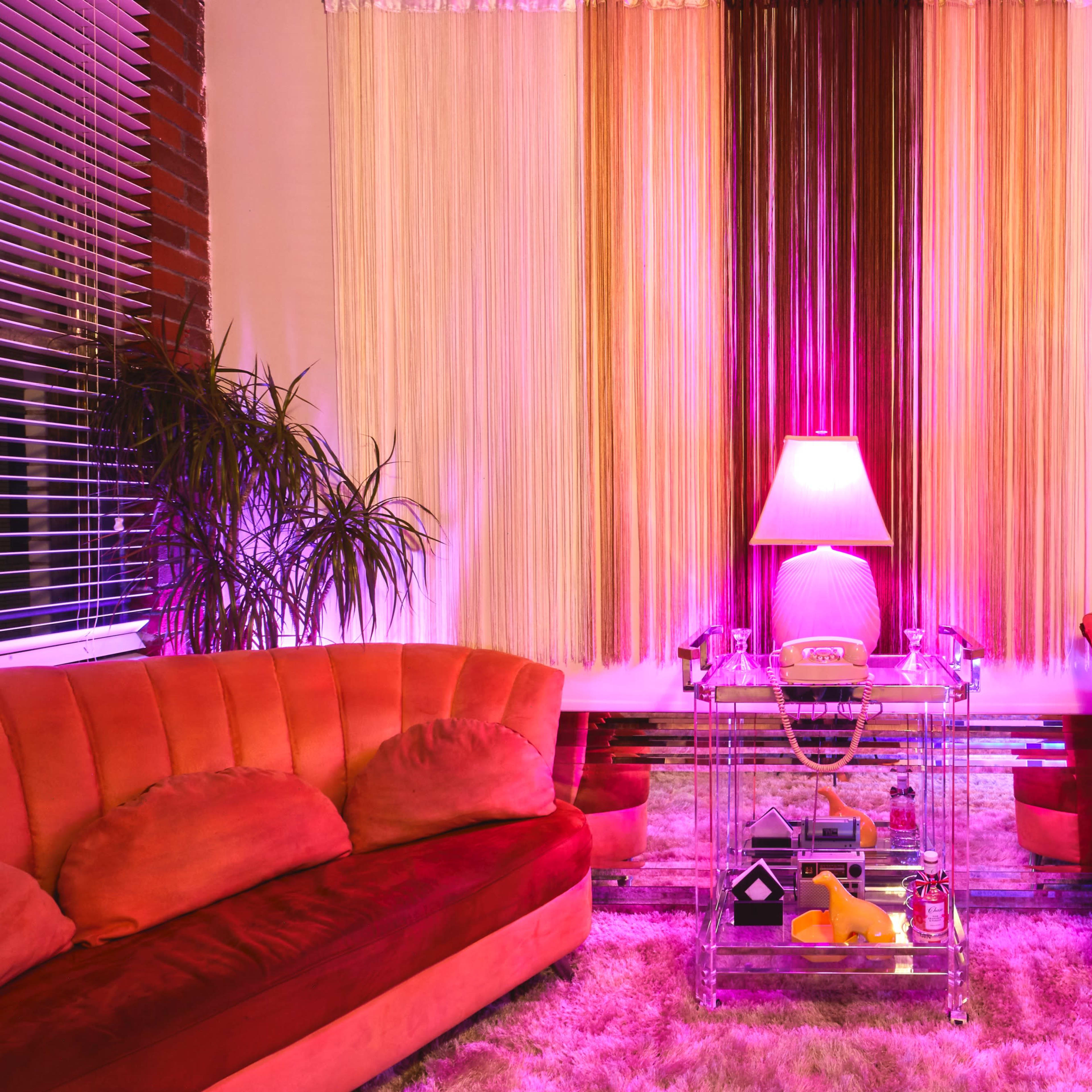 A stylish living room featuring two pink sofas, a glass coffee table with decorative items, and a glowing lamp surrounded by plants and colorful curtains.