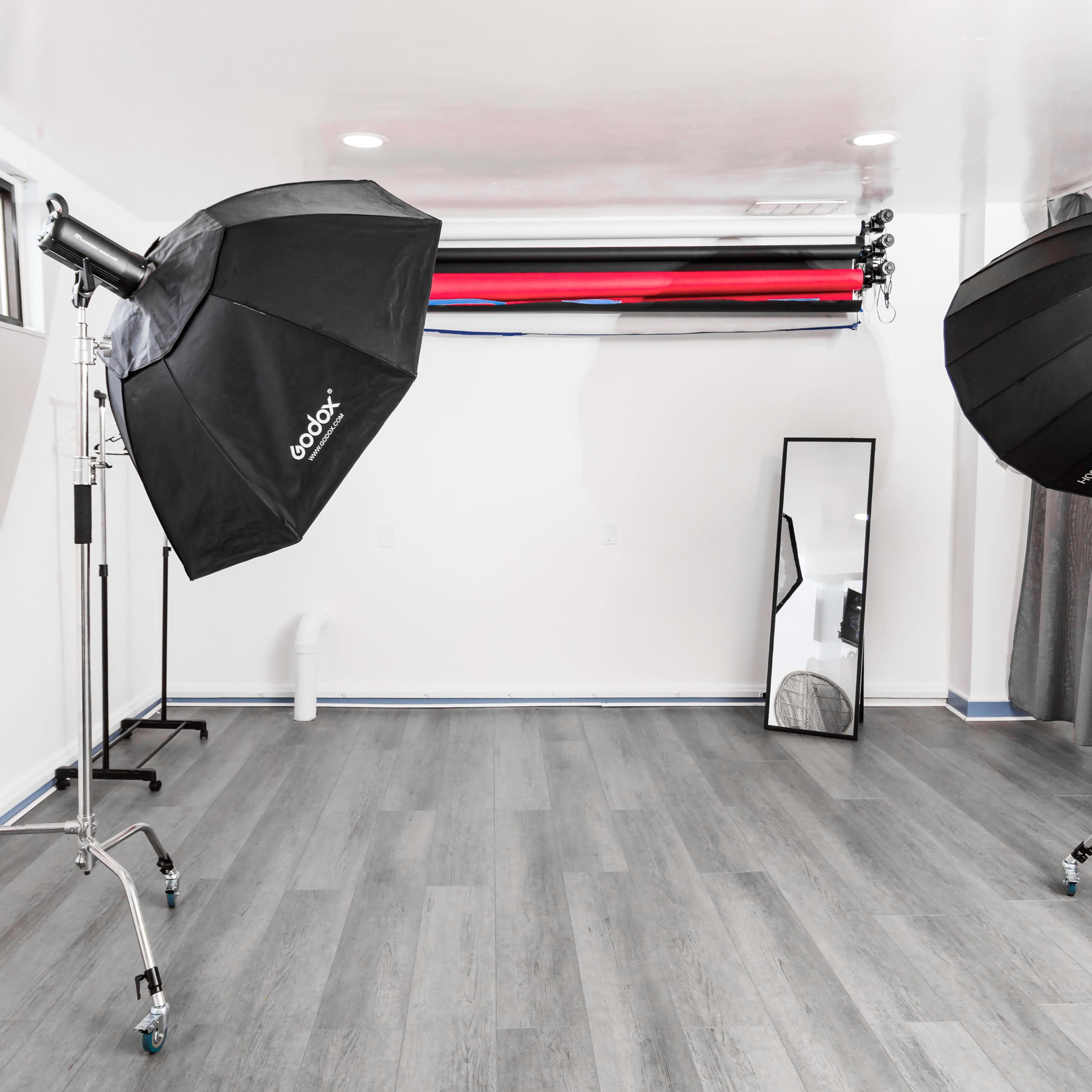 A photography studio with two light softboxes on stands, a large mirror, and a backdrop setup featuring red and black rolls.
