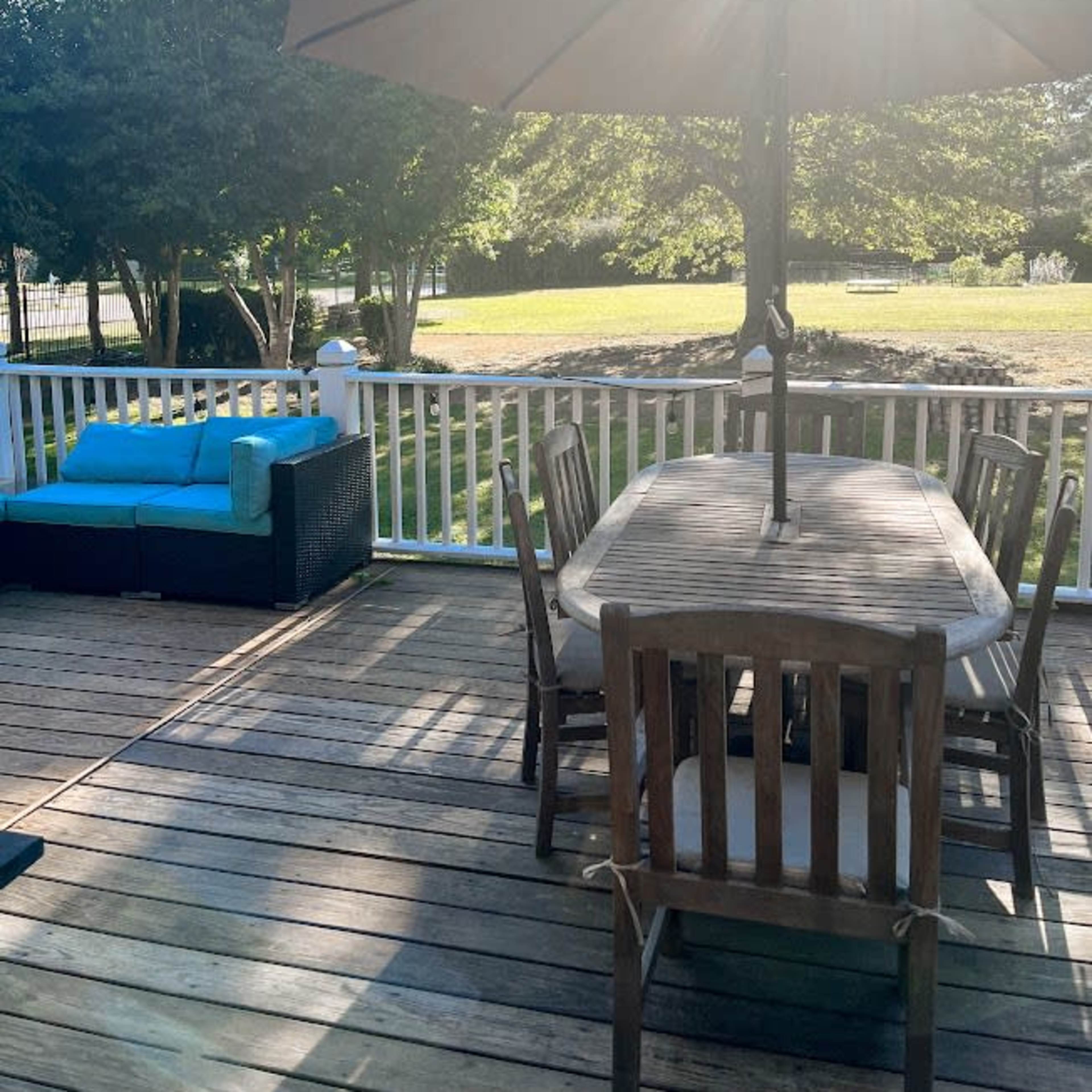 A wooden deck features a dining table with chairs, a lounge area with a blue cushion, and a large umbrella, surrounded by greenery and sunlight.