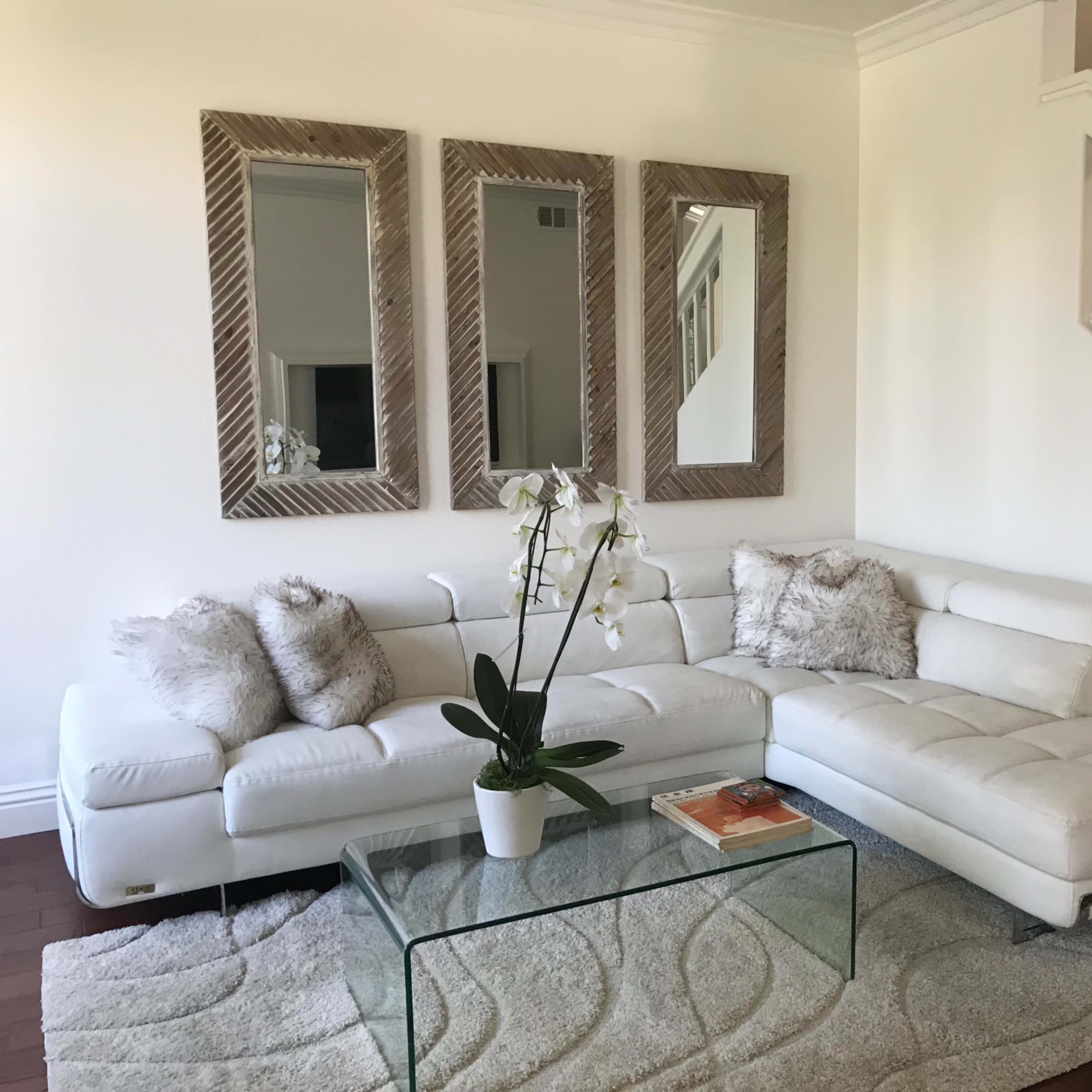 A modern living room with a white sectional sofa, a glass coffee table, and three mirrors mounted on the wall.