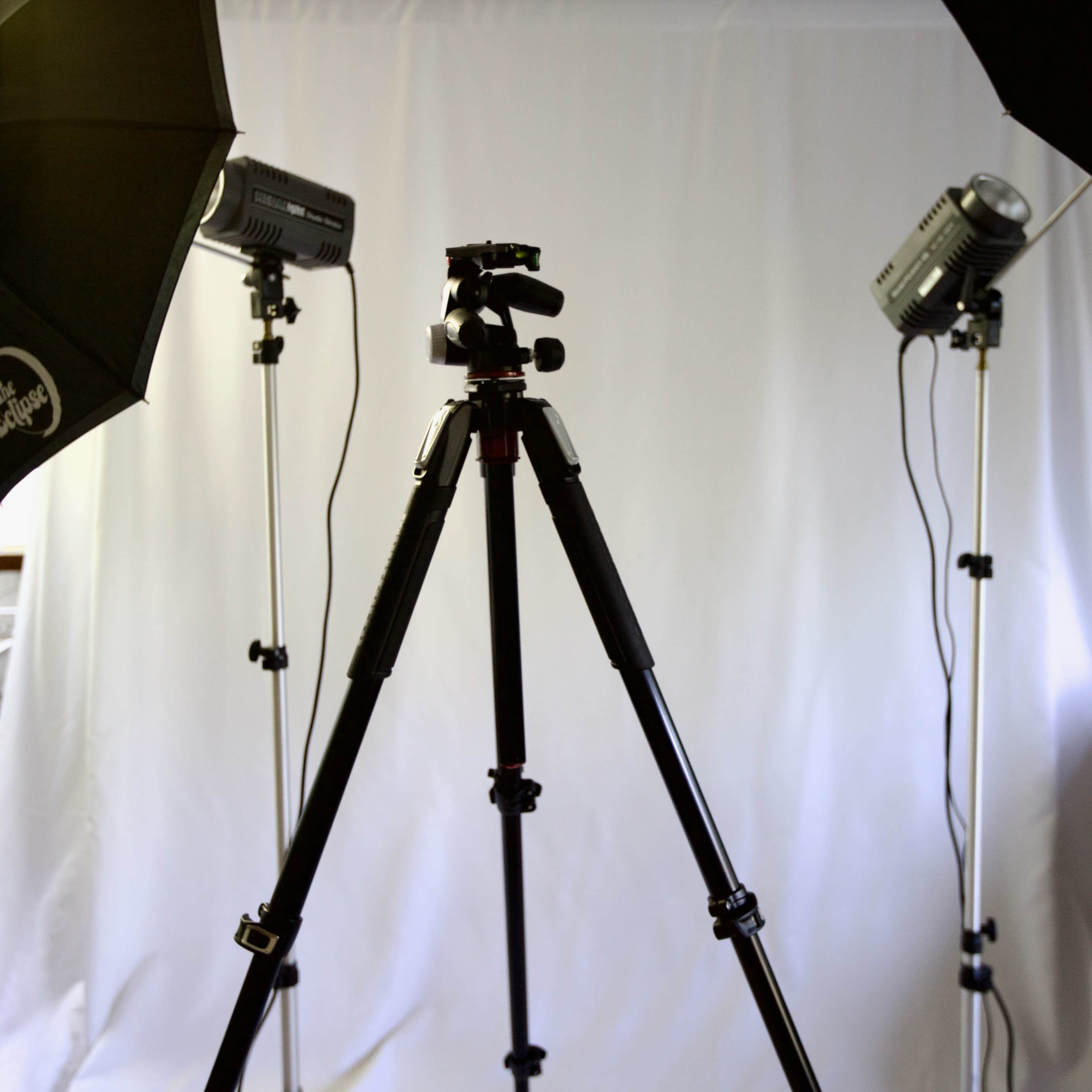 A photography setup with a tripod in the center, flanked by two studio lights and a backdrop.