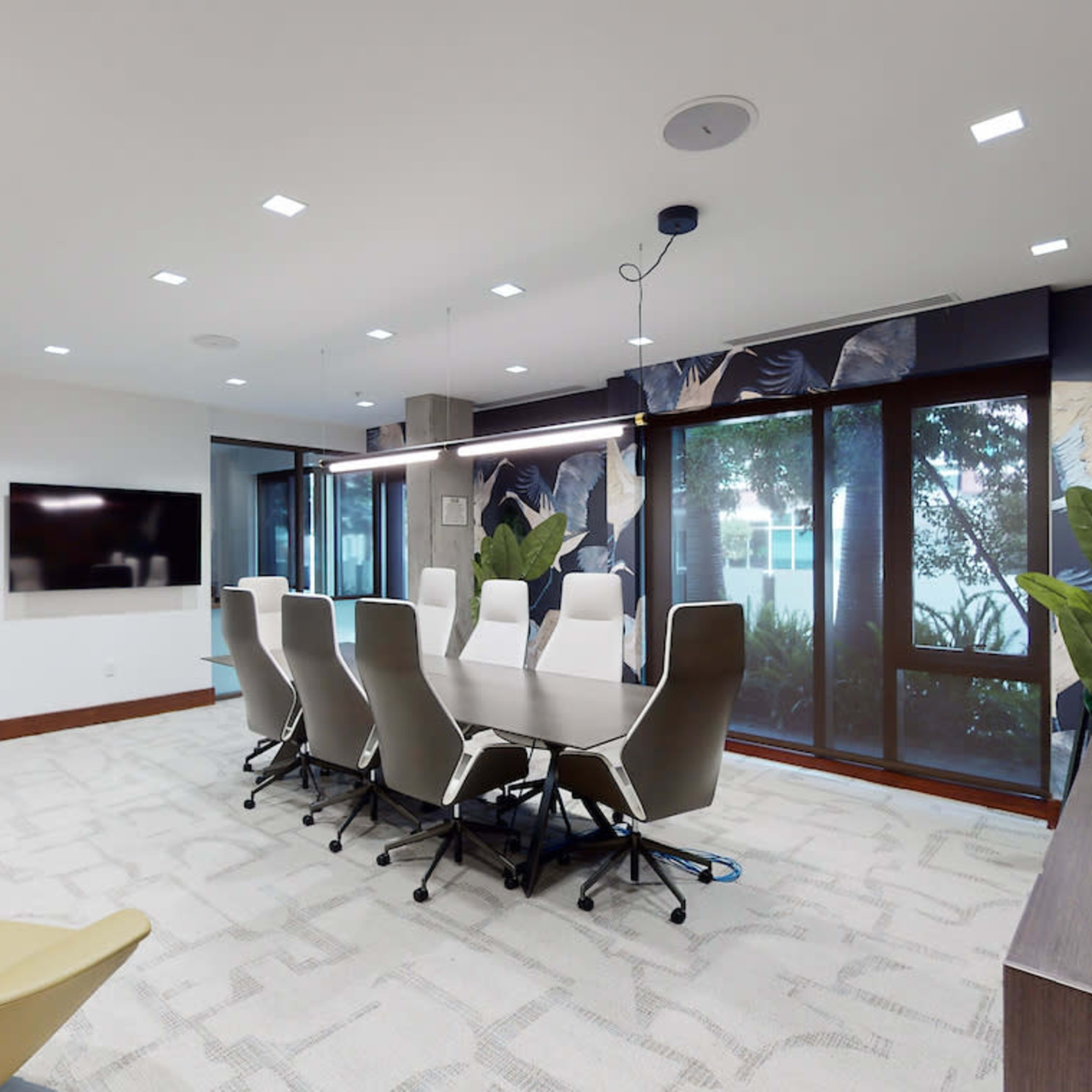 A modern conference room features a long table surrounded by ergonomic chairs, large windows with indoor plants, and a wall-mounted screen.
