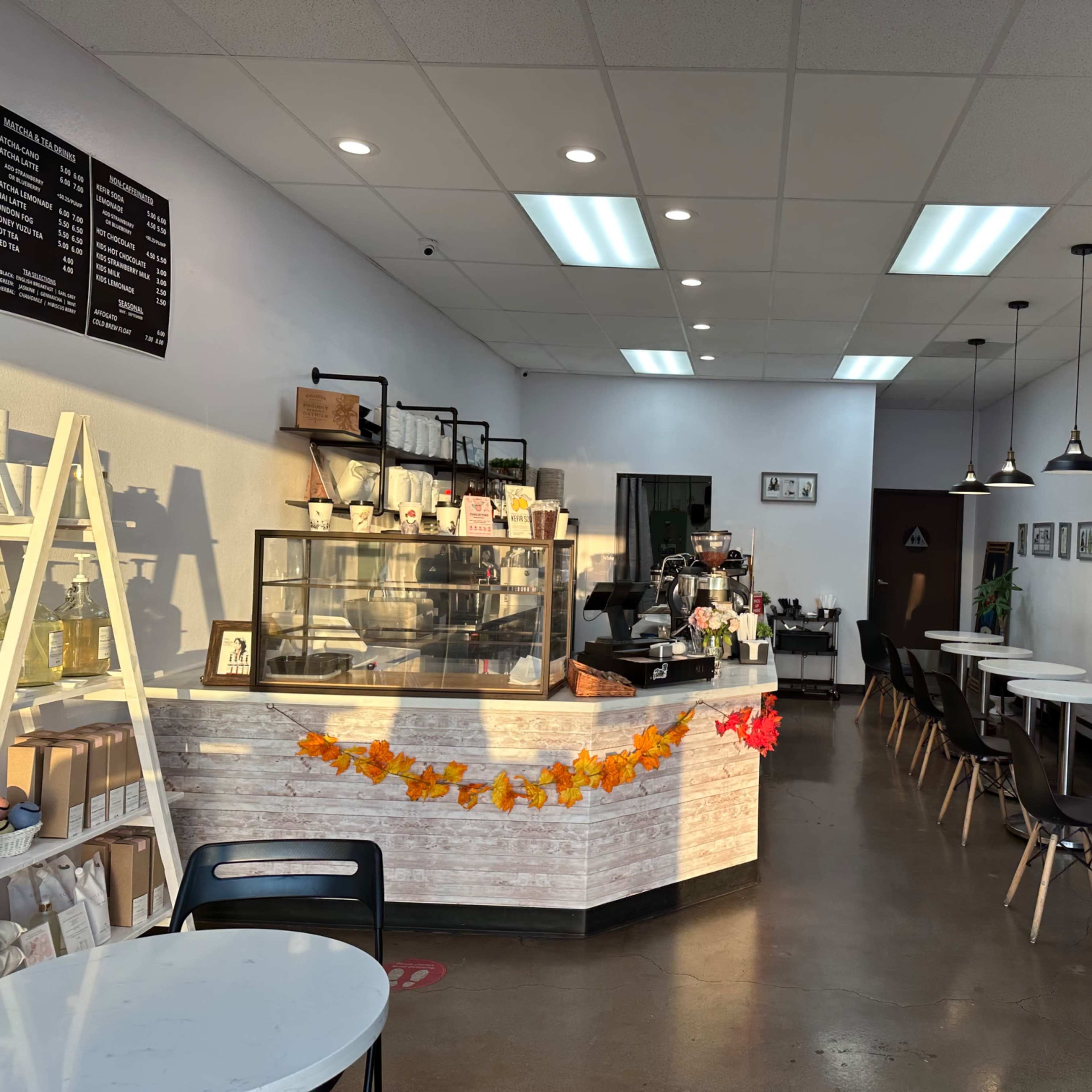 A cozy café interior with a counter displaying beverages, tables arranged along the walls, and framed pictures hanging on the white walls.