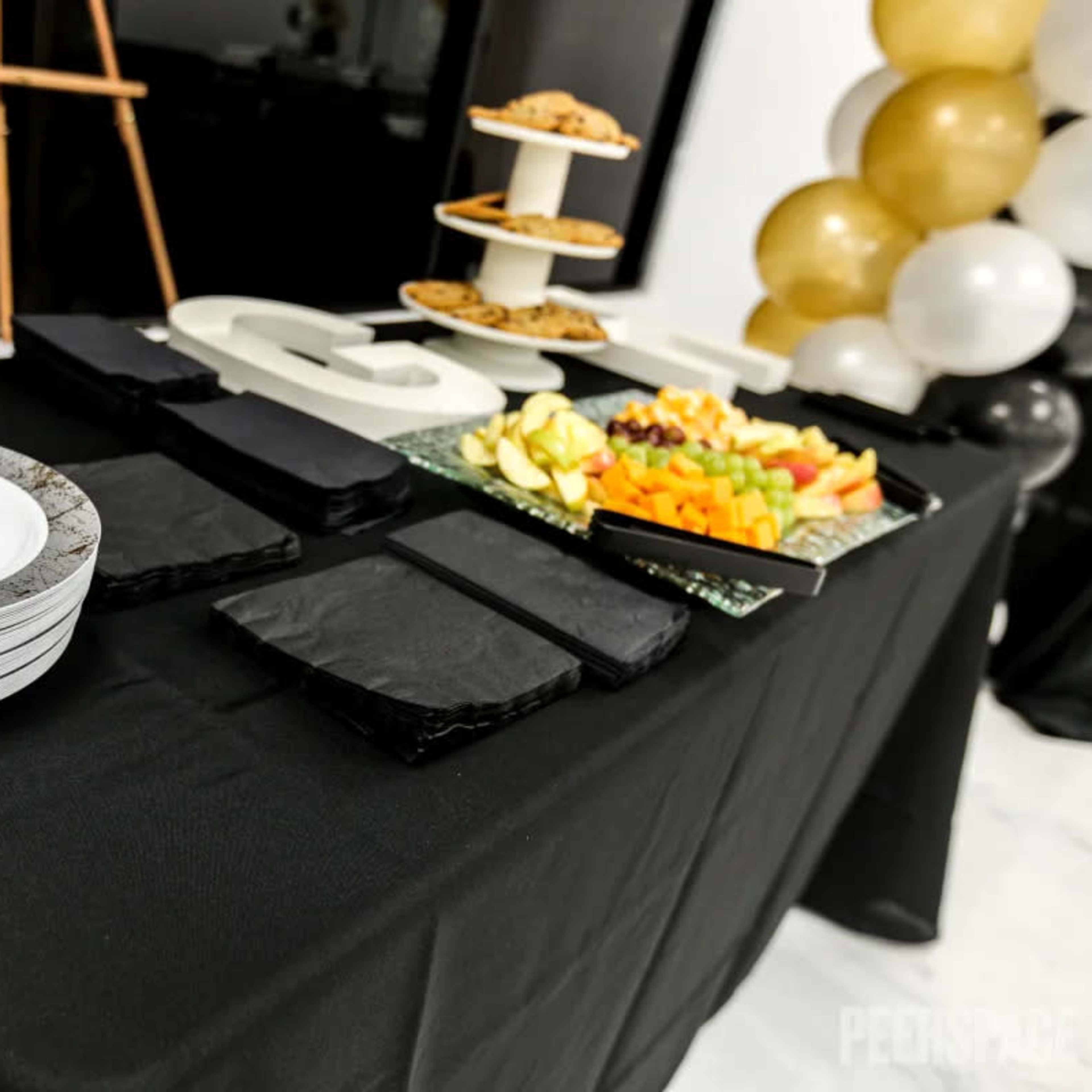 A black table displays plates, utensils, and a variety of fruits and cookies arranged on stands.