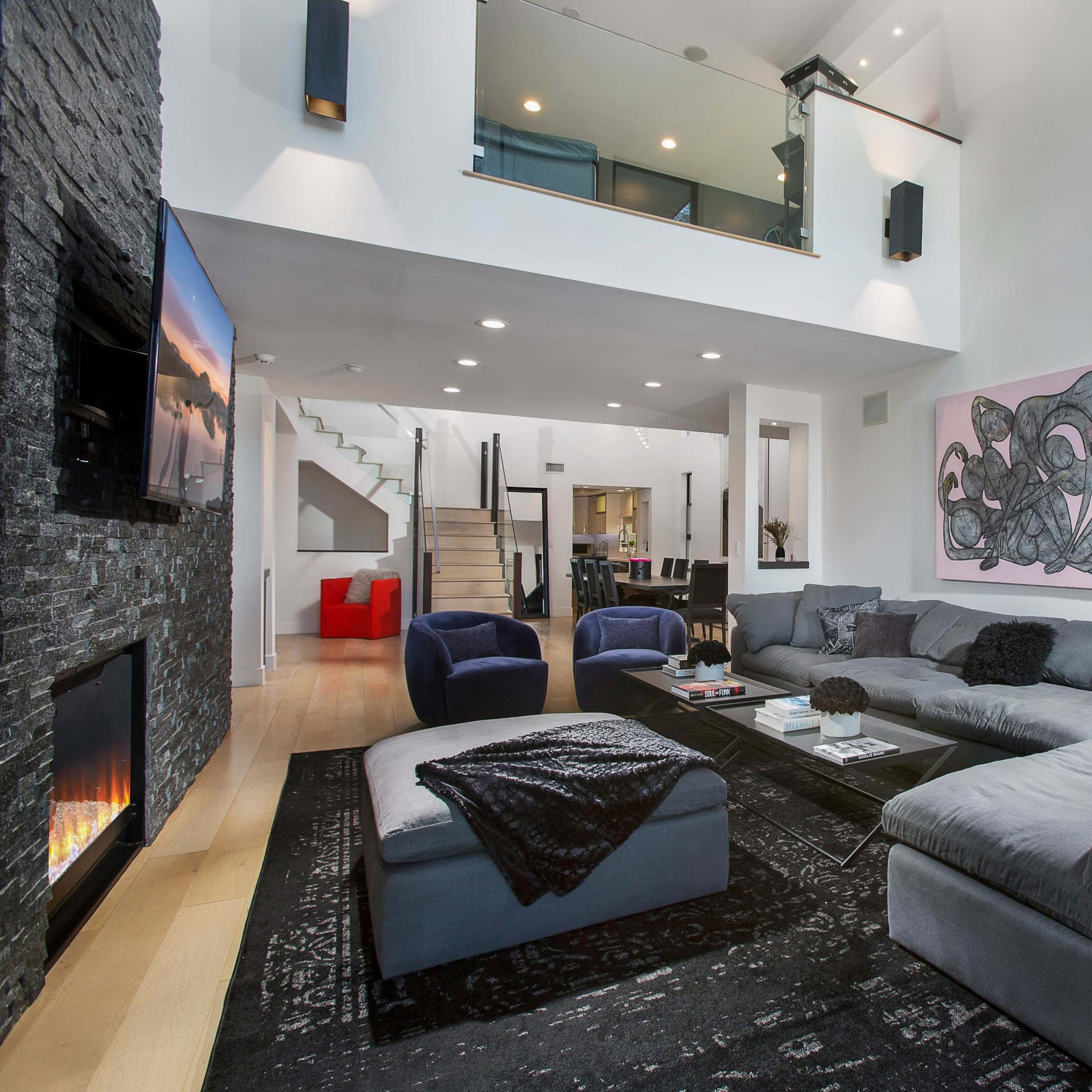 A modern living room features a large sectional sofa, a wall-mounted television, and a prominent artwork, with a staircase leading to a second floor.