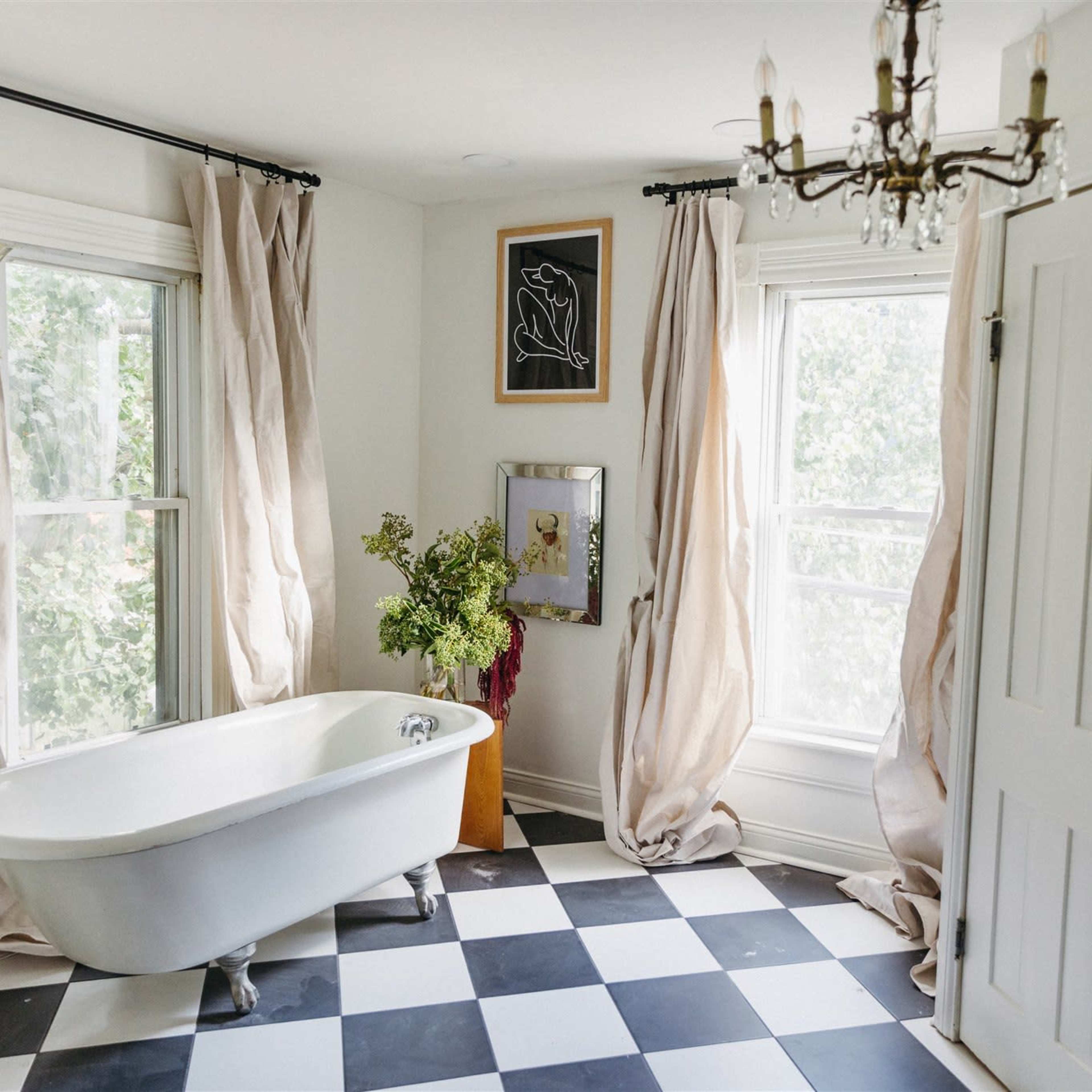 A bathroom with a freestanding bathtub, large windows covered by flowing curtains, tiled black and white flooring, and a closet with double doors.
