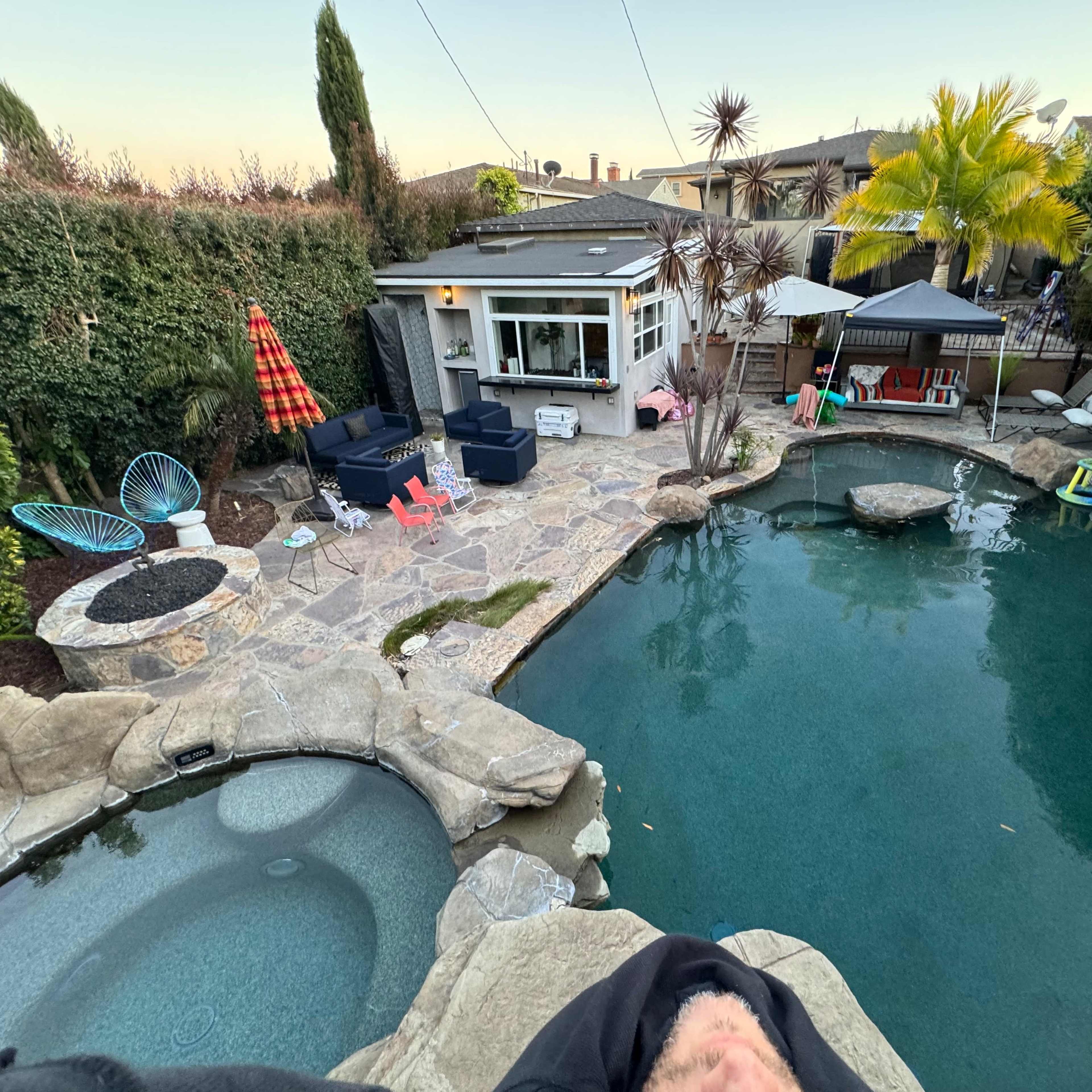 A backyard with a swimming pool surrounded by stone decking, lounge chairs, a fire pit, and tropical plants.