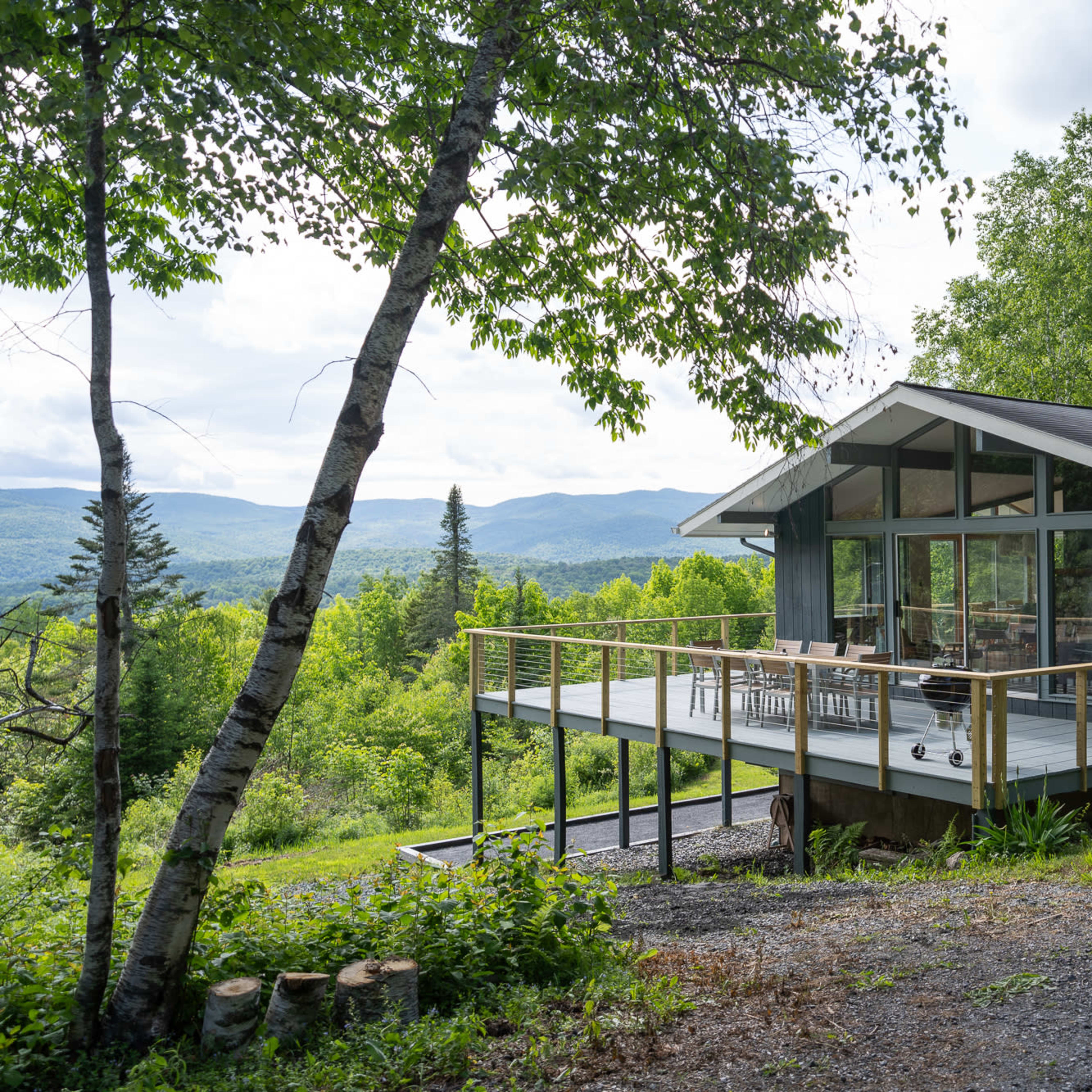 A modern cabin with a large deck overlooks a lush green landscape and distant mountains.