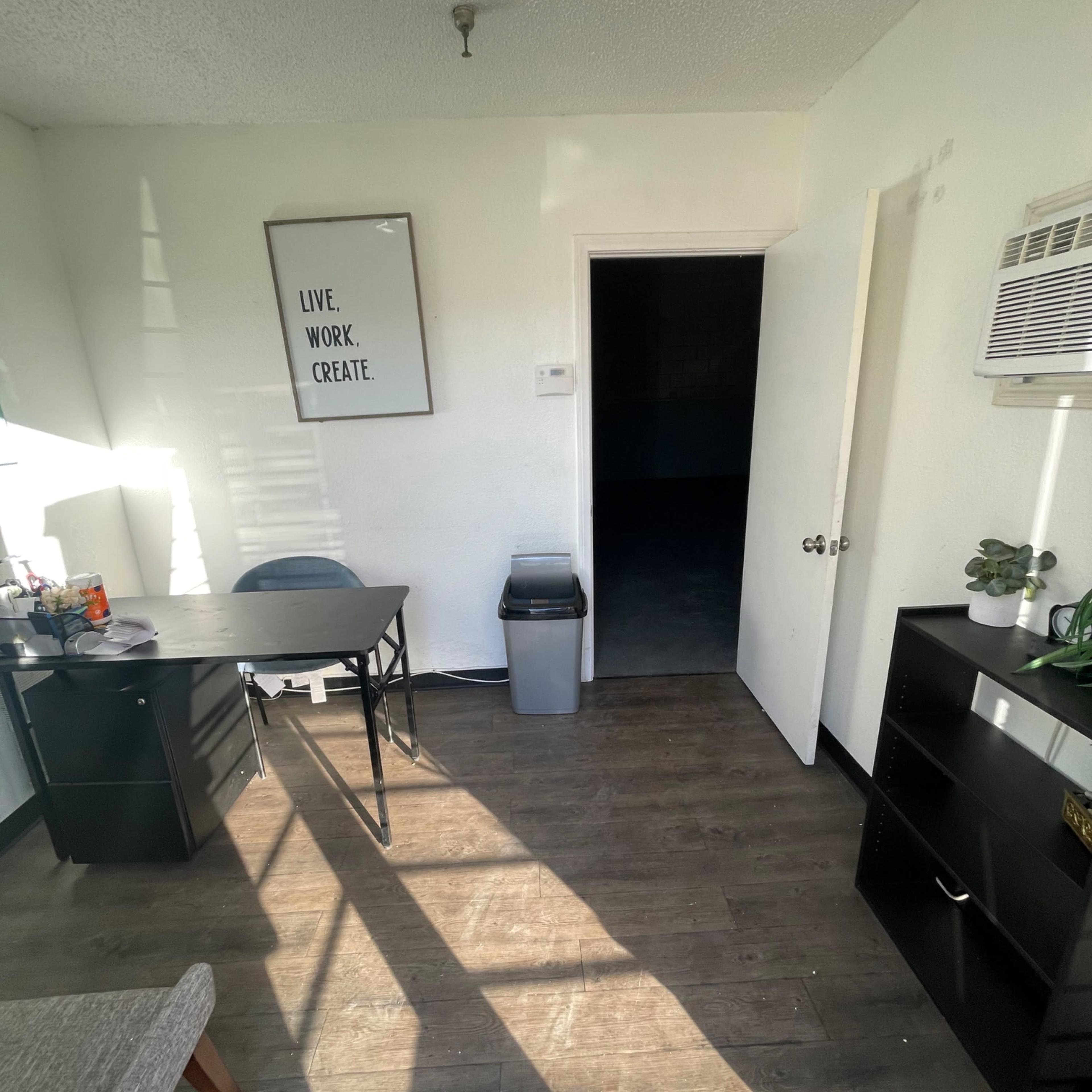 A small office space featuring a desk, a chair, a trash can, and a door leading to a dark room.