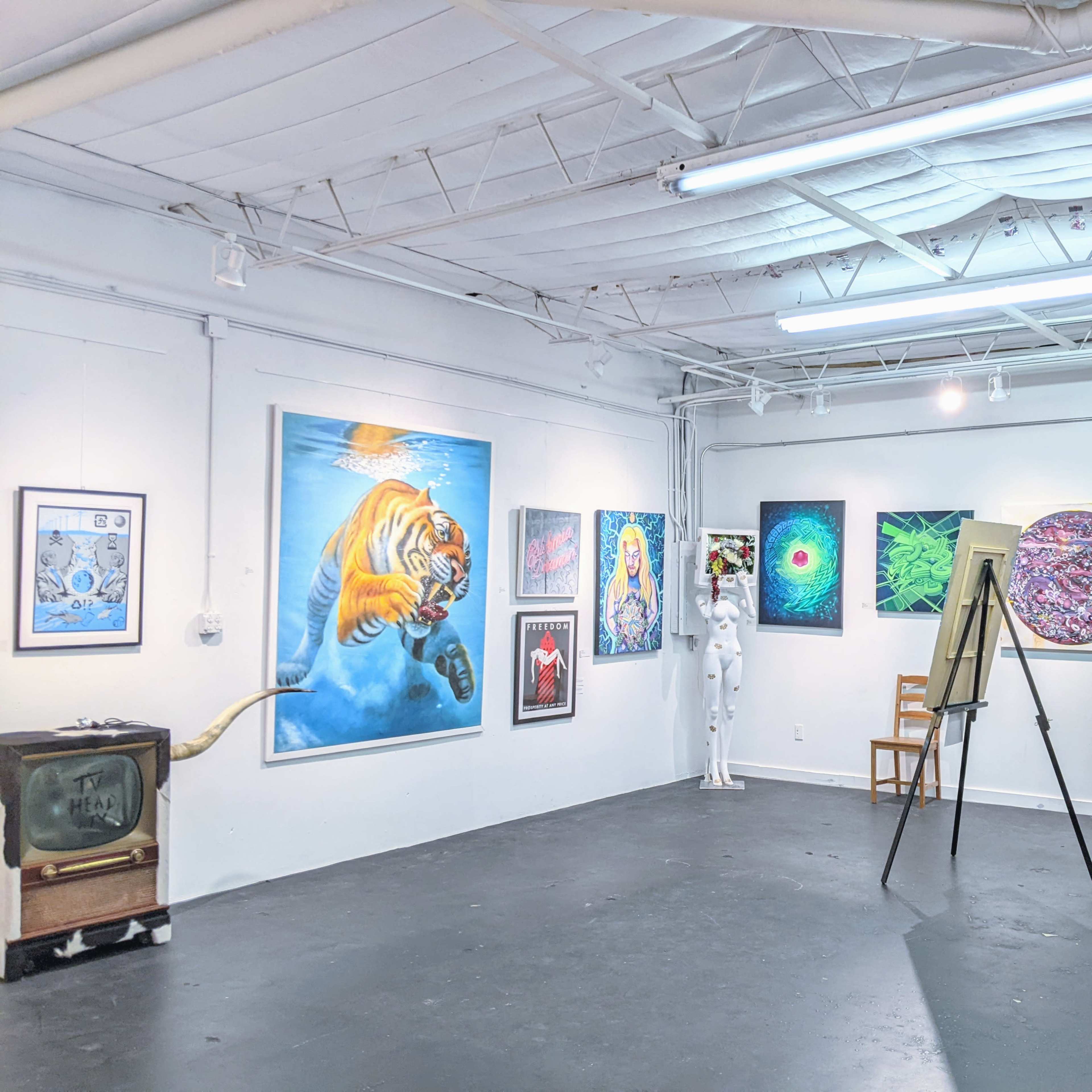 An art gallery with various colorful paintings on white walls, a black display board, and a mannequin in a white suit standing in the corner.