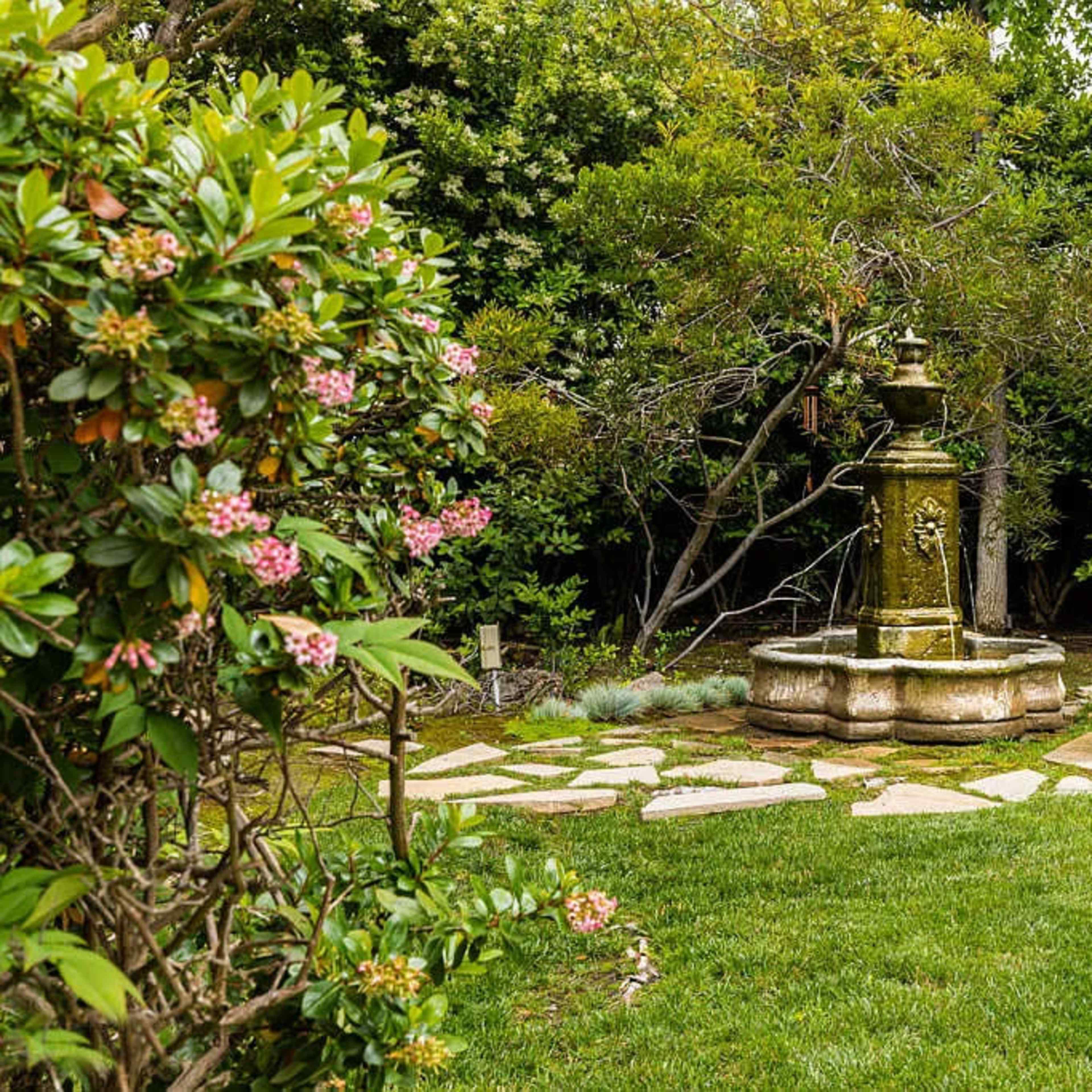 A stone fountain surrounded by a manicured lawn and lush greenery creates a tranquil garden scene.
