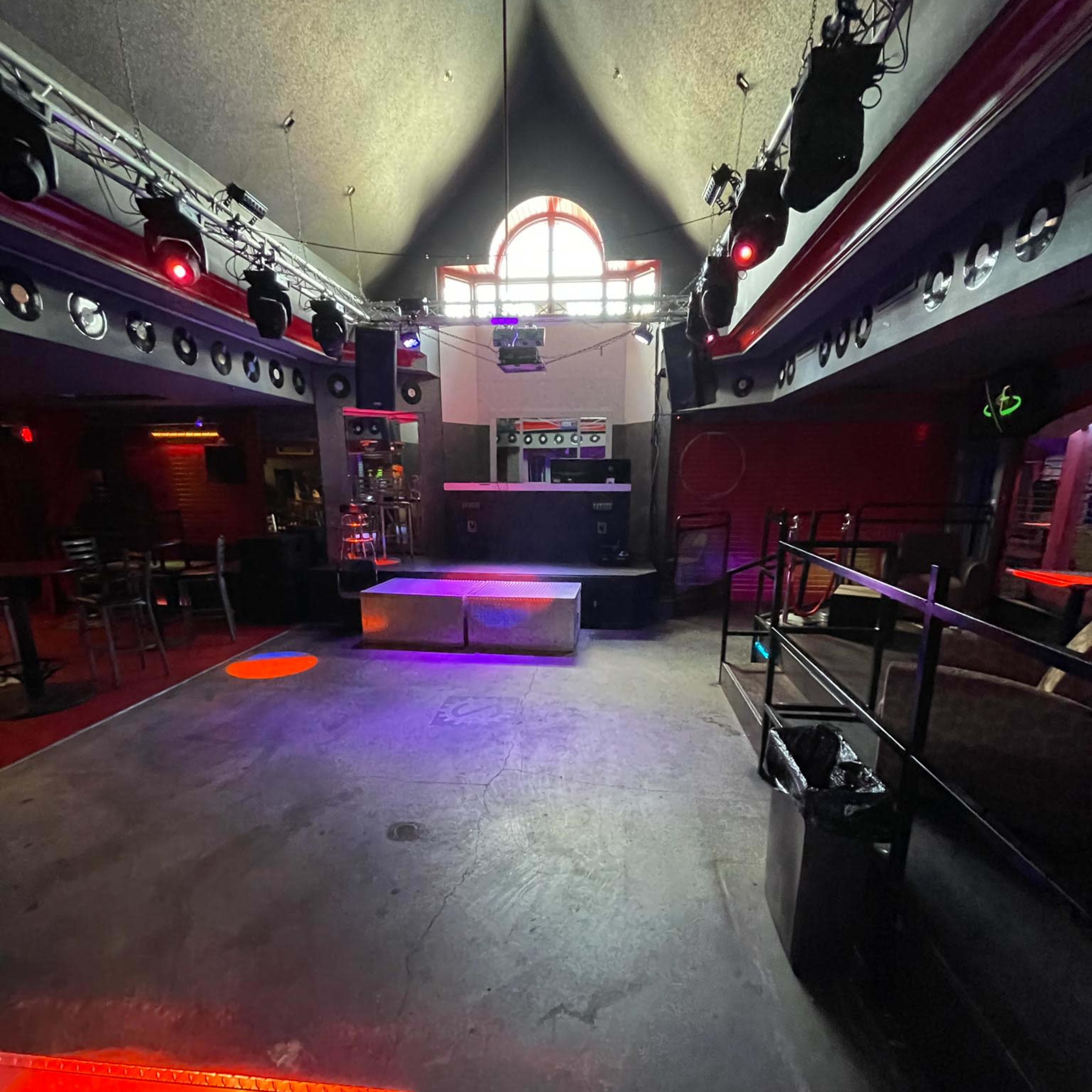An empty nightclub interior with a DJ booth at the back, illuminated by red and purple lights.