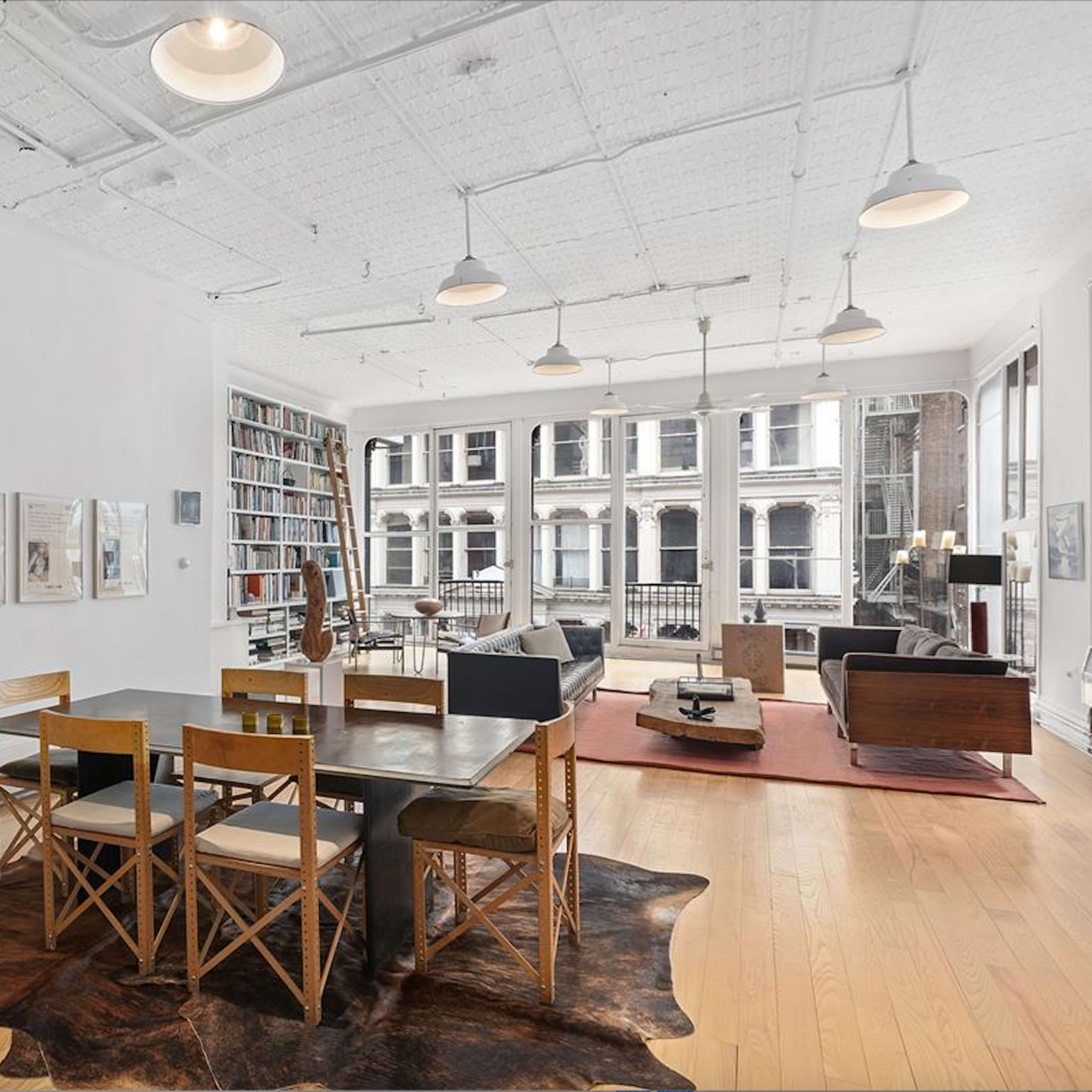 A modern, open living space with a dining area featuring a black table and wooden chairs, large windows overlooking a cityscape, and a collection of bookshelves against the wall.