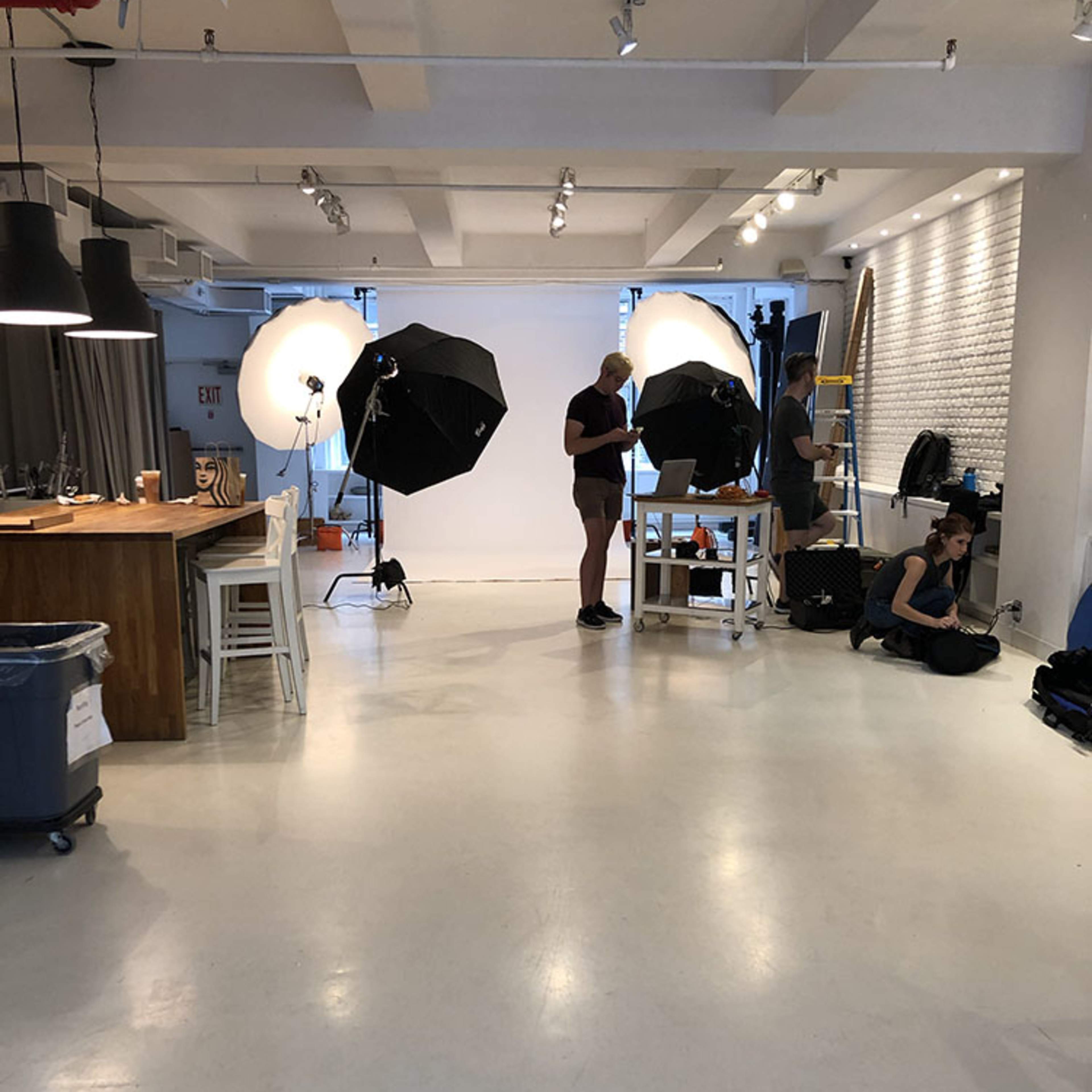 A photography studio is set up with two large light umbrellas, a backdrop, and individuals arranging equipment.