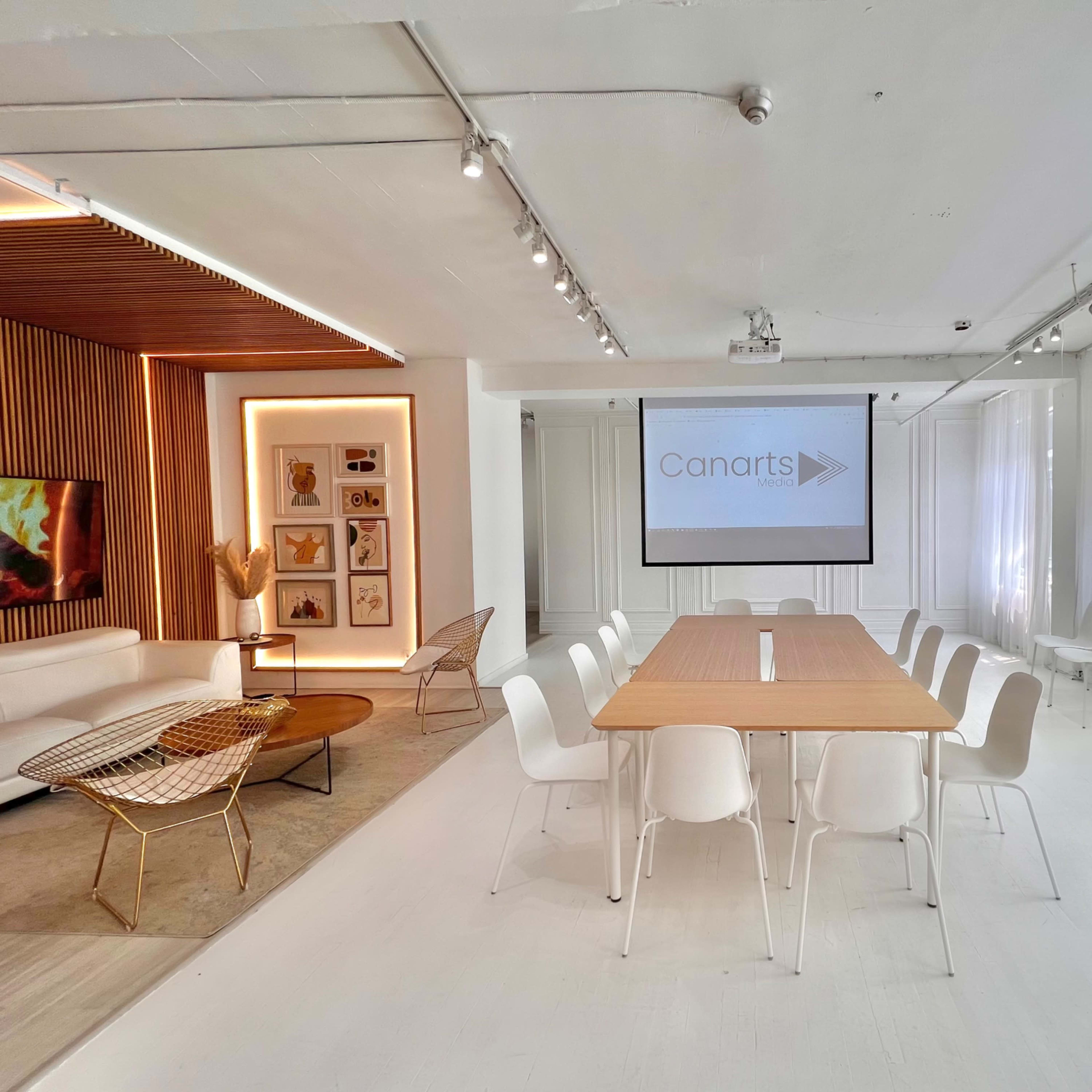 A modern conference room features a large wooden table surrounded by white chairs, with a projector screen displaying "Canarts" on one wall.