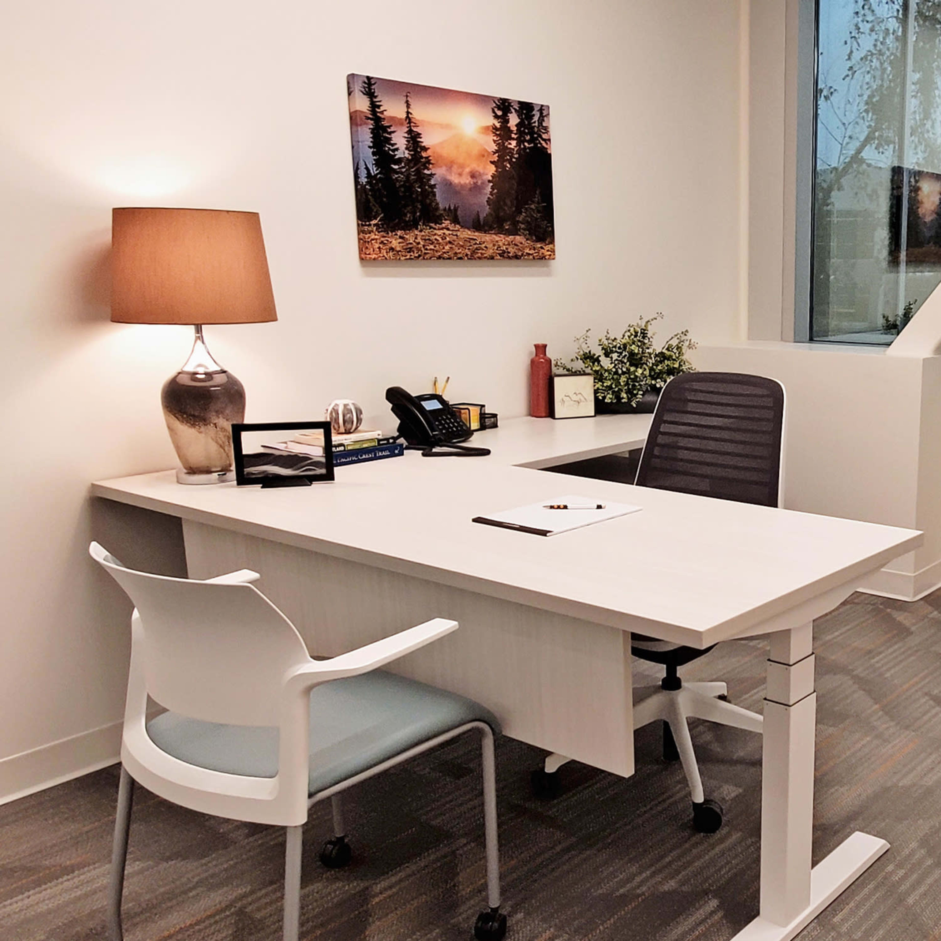 A modern office space featuring a desk, a chair, a lamp, and large windows with natural light.