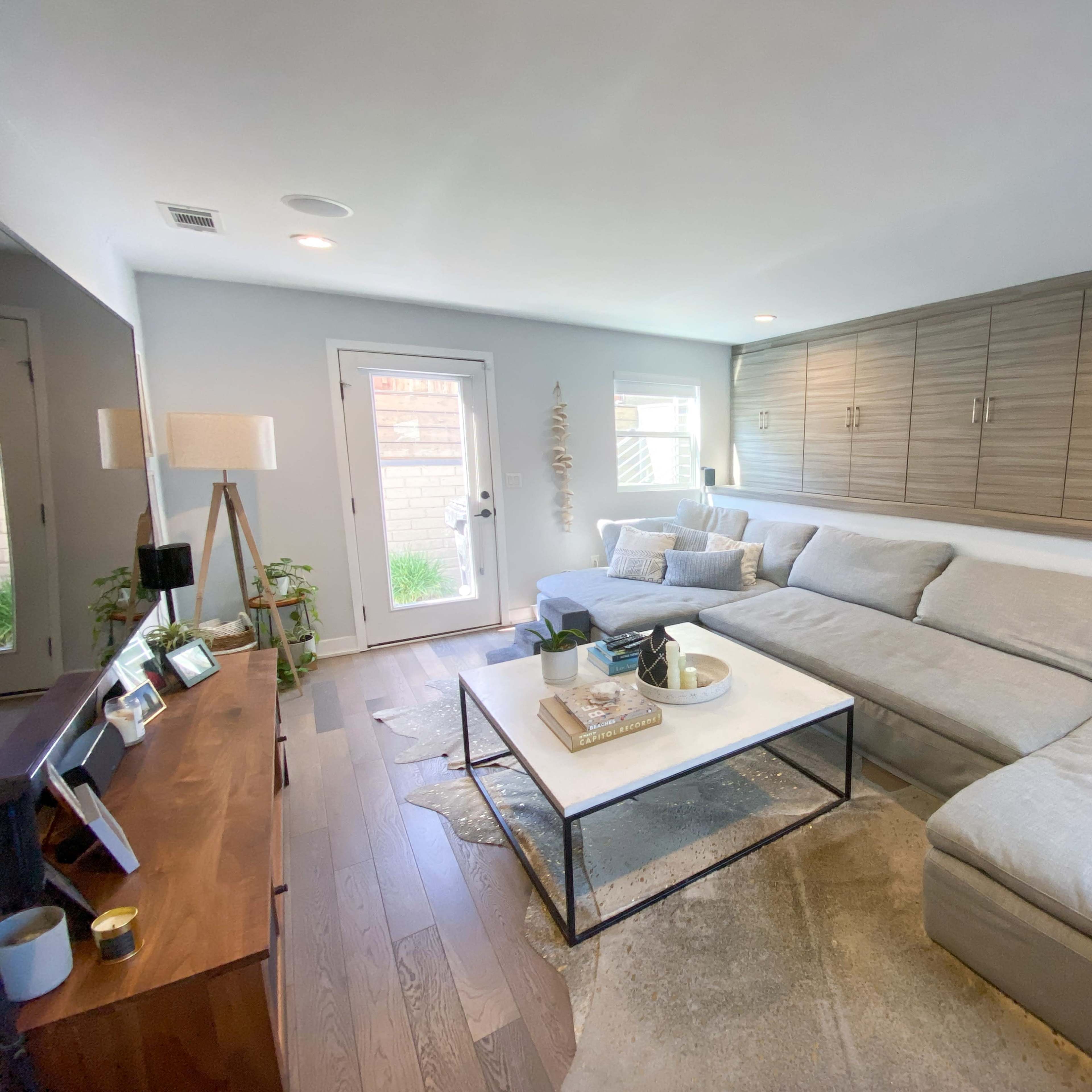 A modern living room featuring a large gray sectional sofa, a coffee table on a light rug, and a door leading to a garden outside.