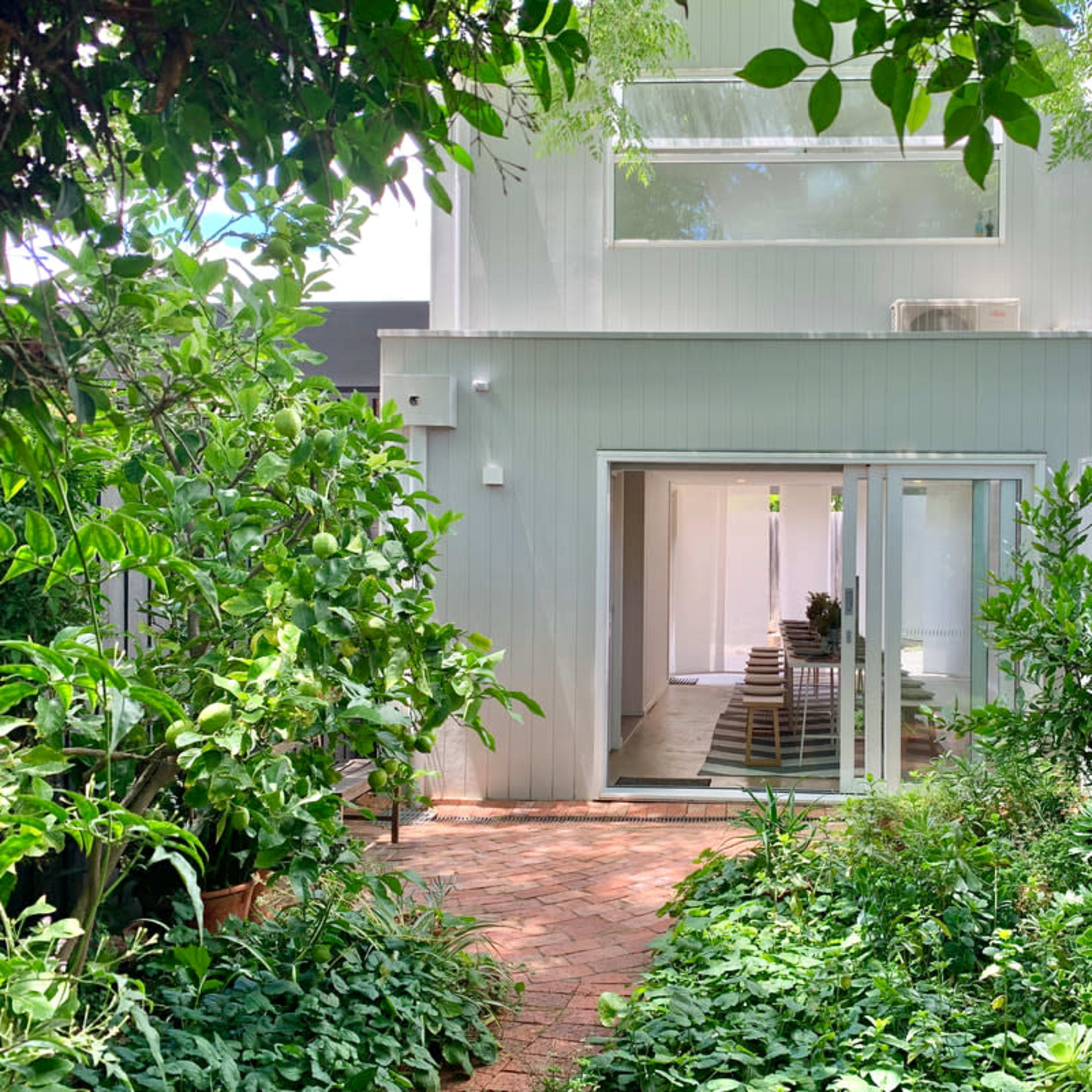 A modern, two-story house with large glass doors is surrounded by greenery and a brick pathway.
