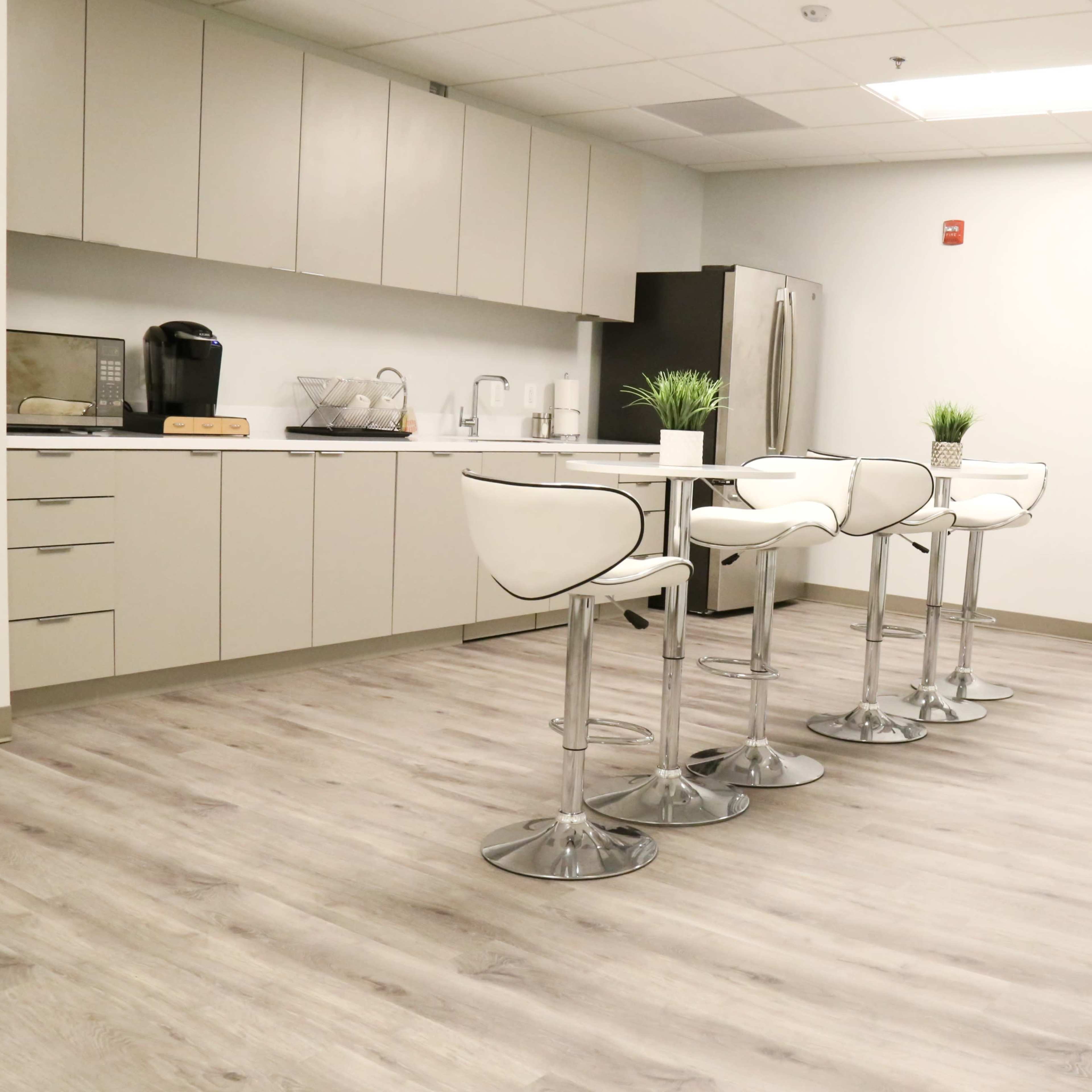 A modern kitchen area with a bar table and four white stools, complemented by cabinets, appliances, and a large window.