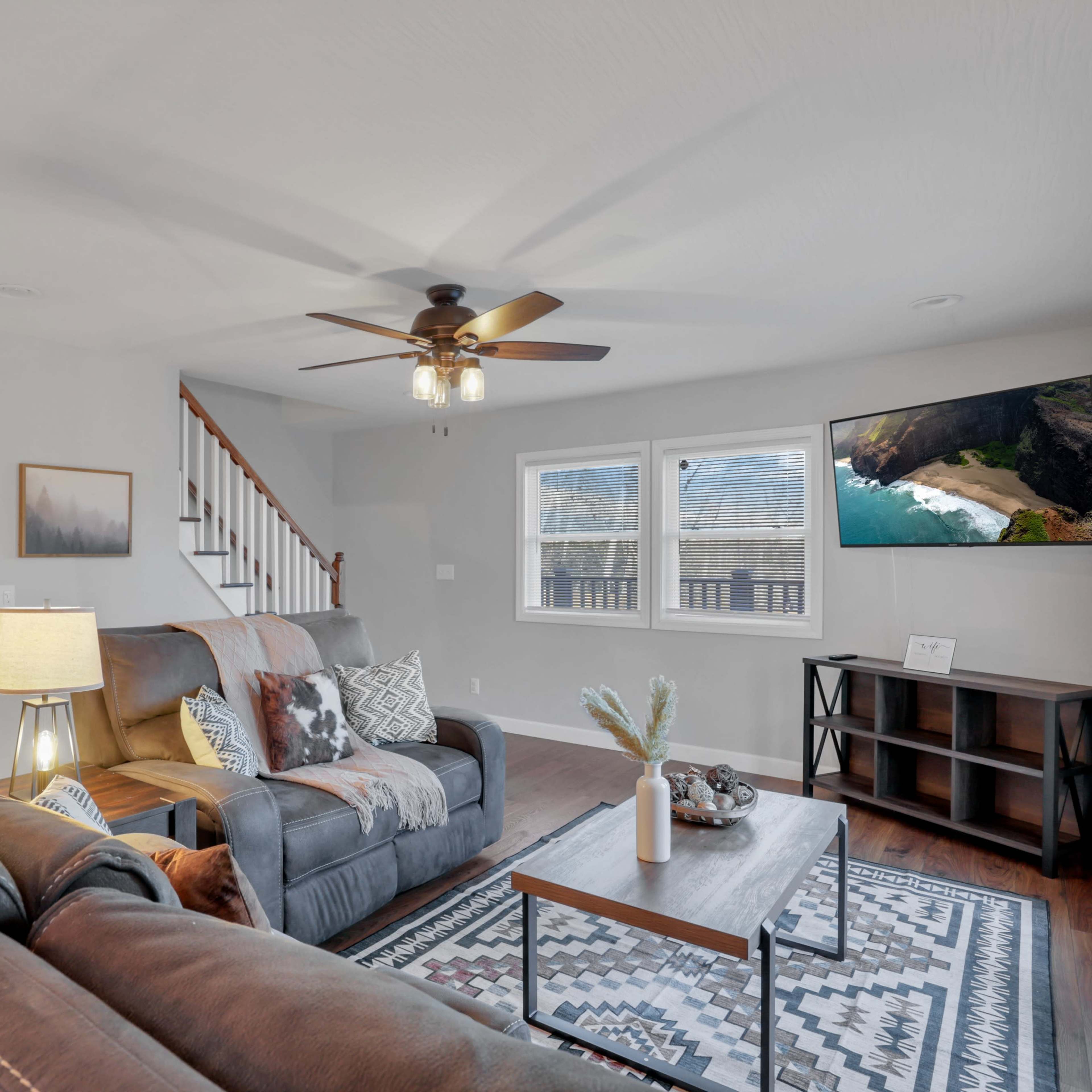 The living room features a sofa and armchair arranged around a coffee table, with a ceiling fan above and windows providing natural light.