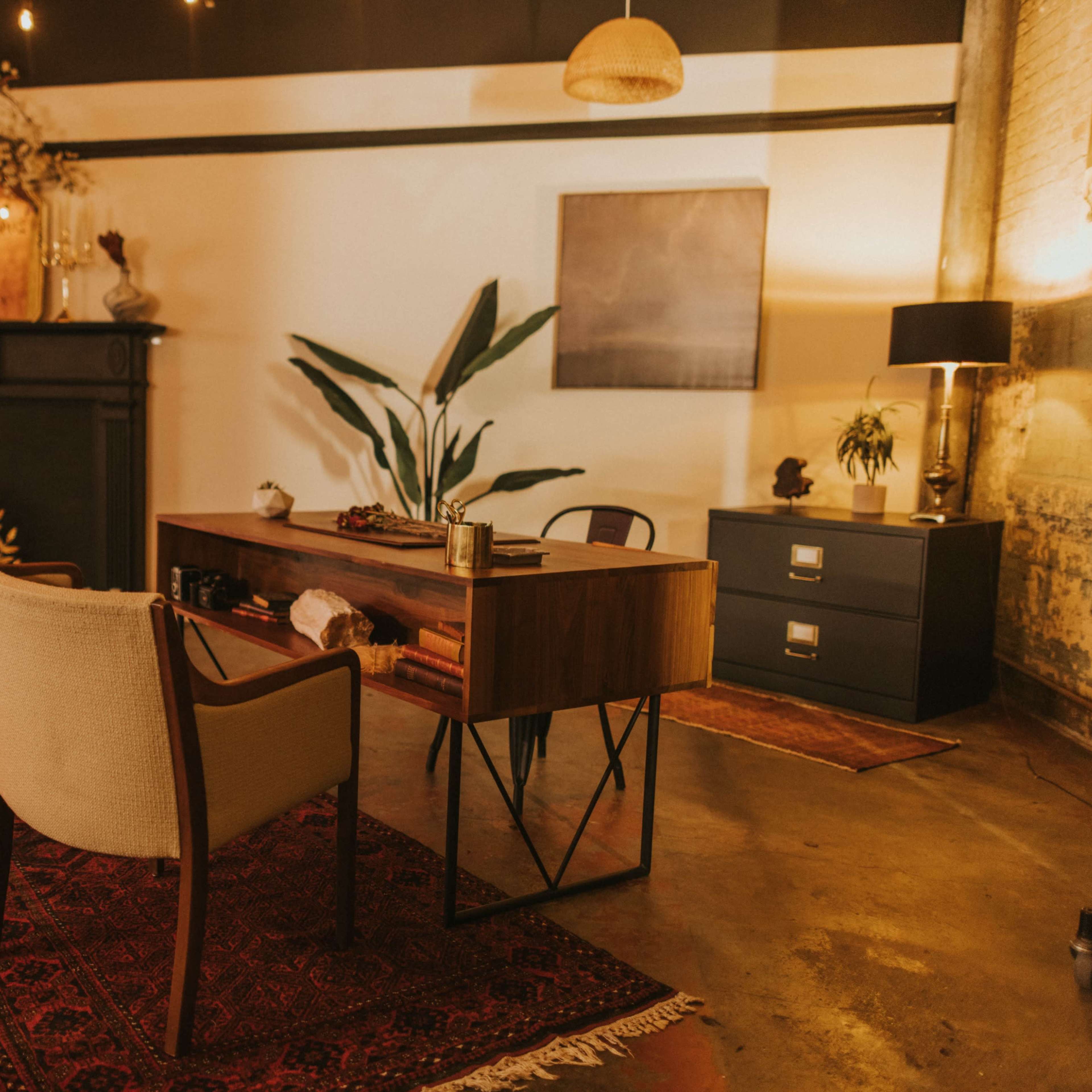 An elegantly styled office space featuring a wooden desk with two cream-colored chairs, a decorative plant, and a dark-colored cabinet against a textured wall.