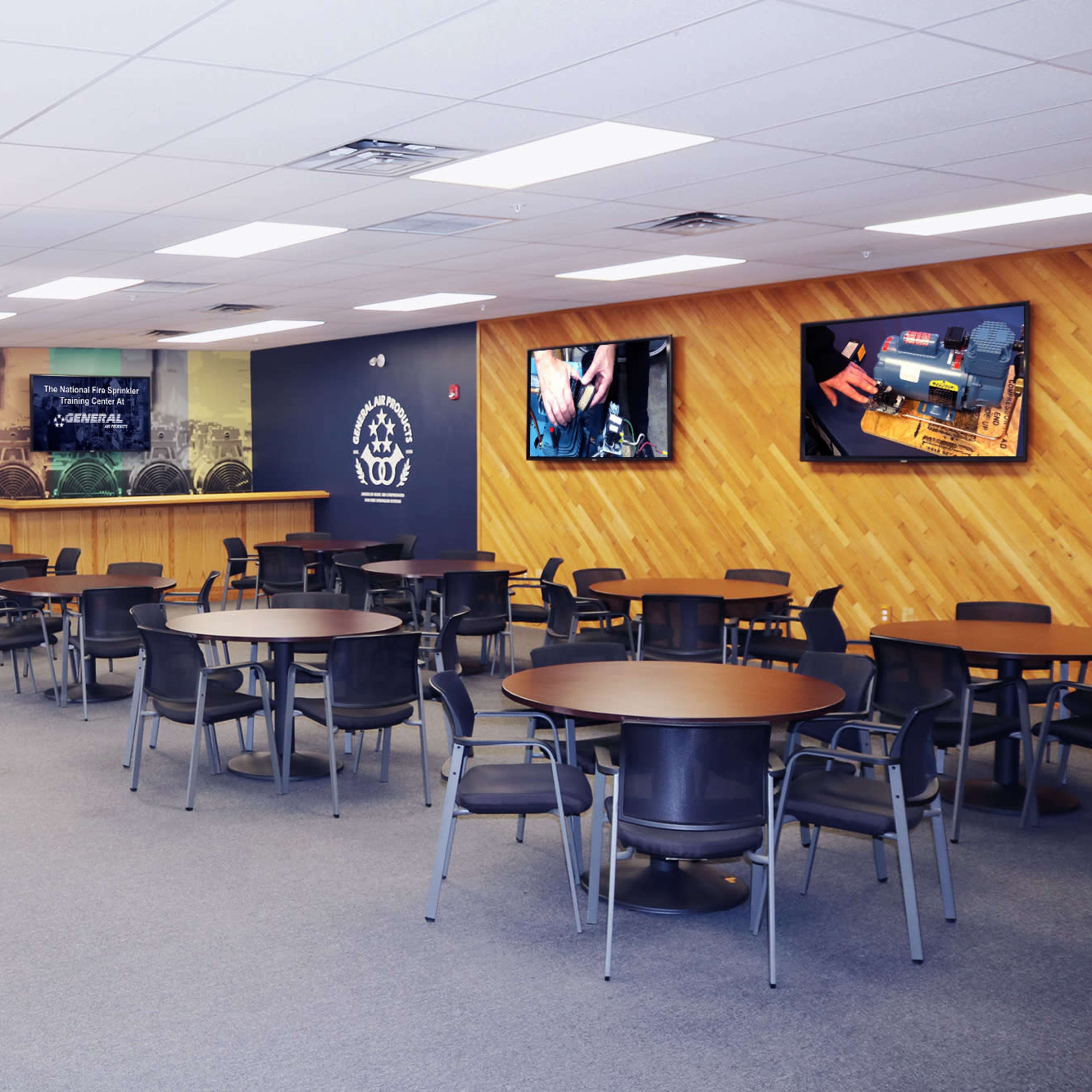 A spacious training room with multiple tables and chairs arranged for groups, along with two large screens displaying technical content on the walls.