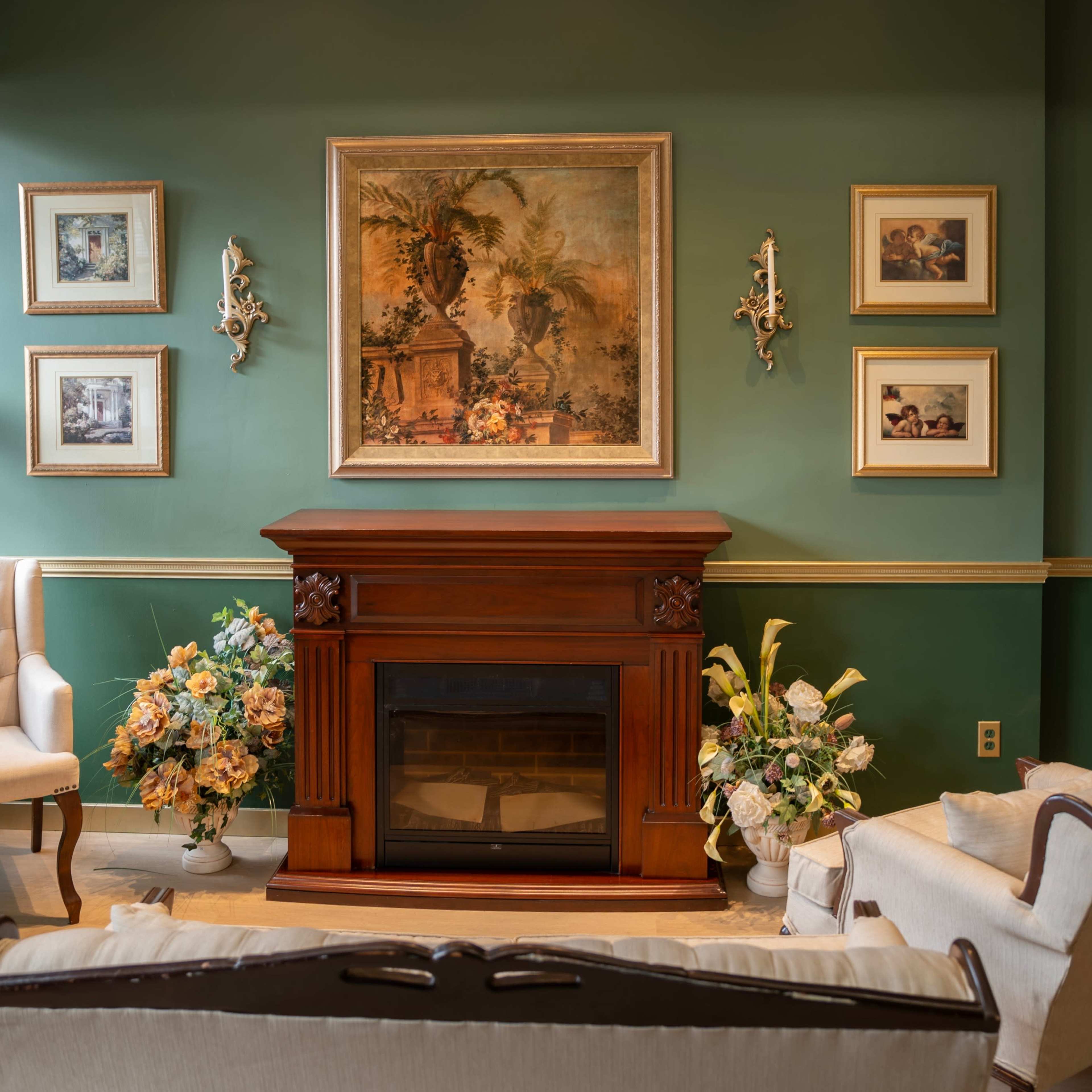 A cozy living room with a fireplace, decorated walls, and floral arrangements beside two upholstered chairs.